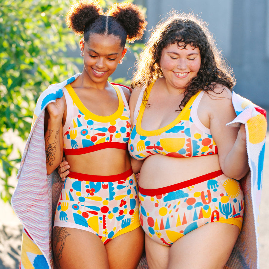 Two models smiling and jumping together wearing sets of Bralettes and underwear in Balance by Lisa Congdon print: geometric shapes in red, yellow, pink, and blue colors.