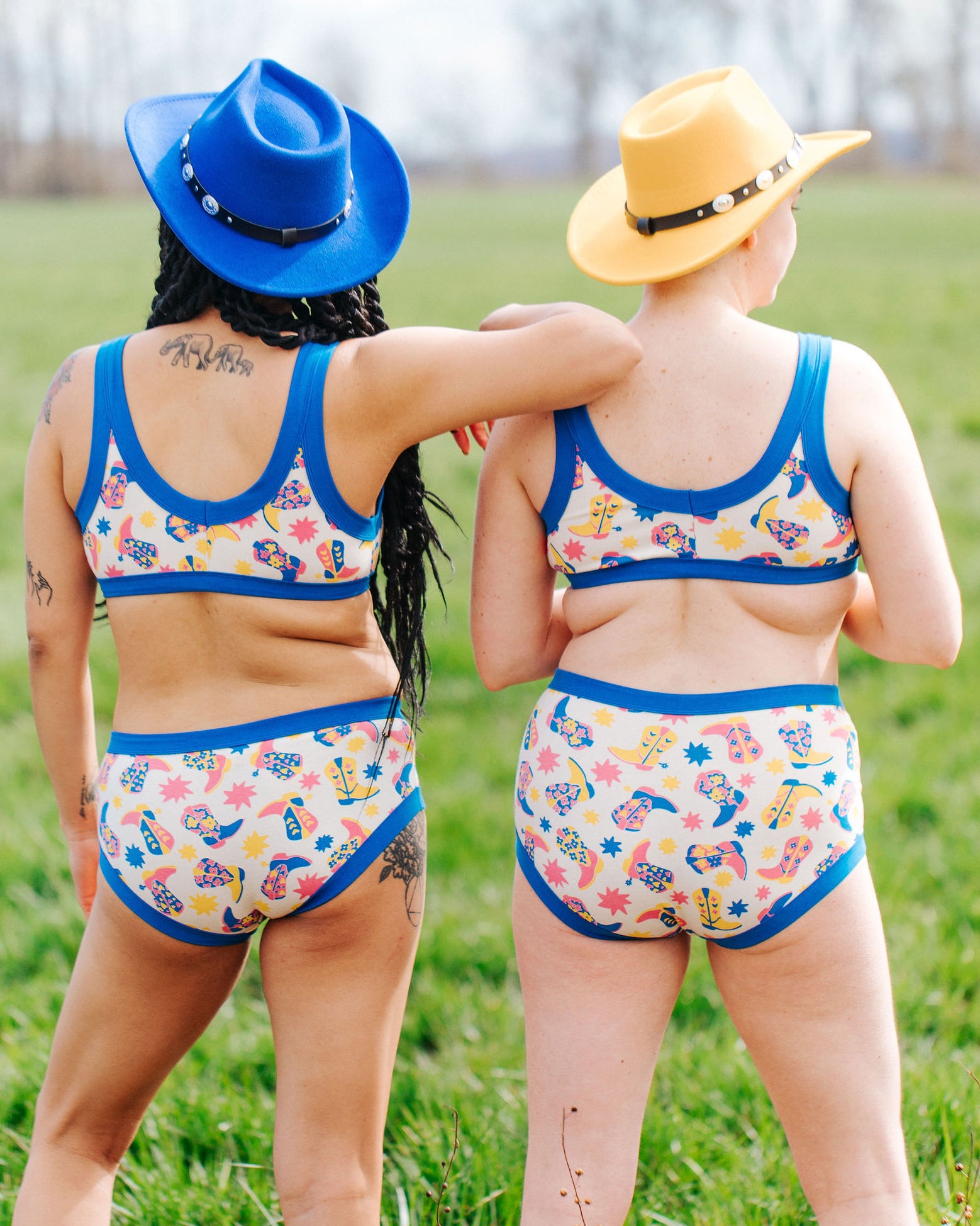 The back of two models standing in a field wearing Thunderpants Bralette and underwear sets in Boot Scootin' - fun boots in pink, yellow, and blue.