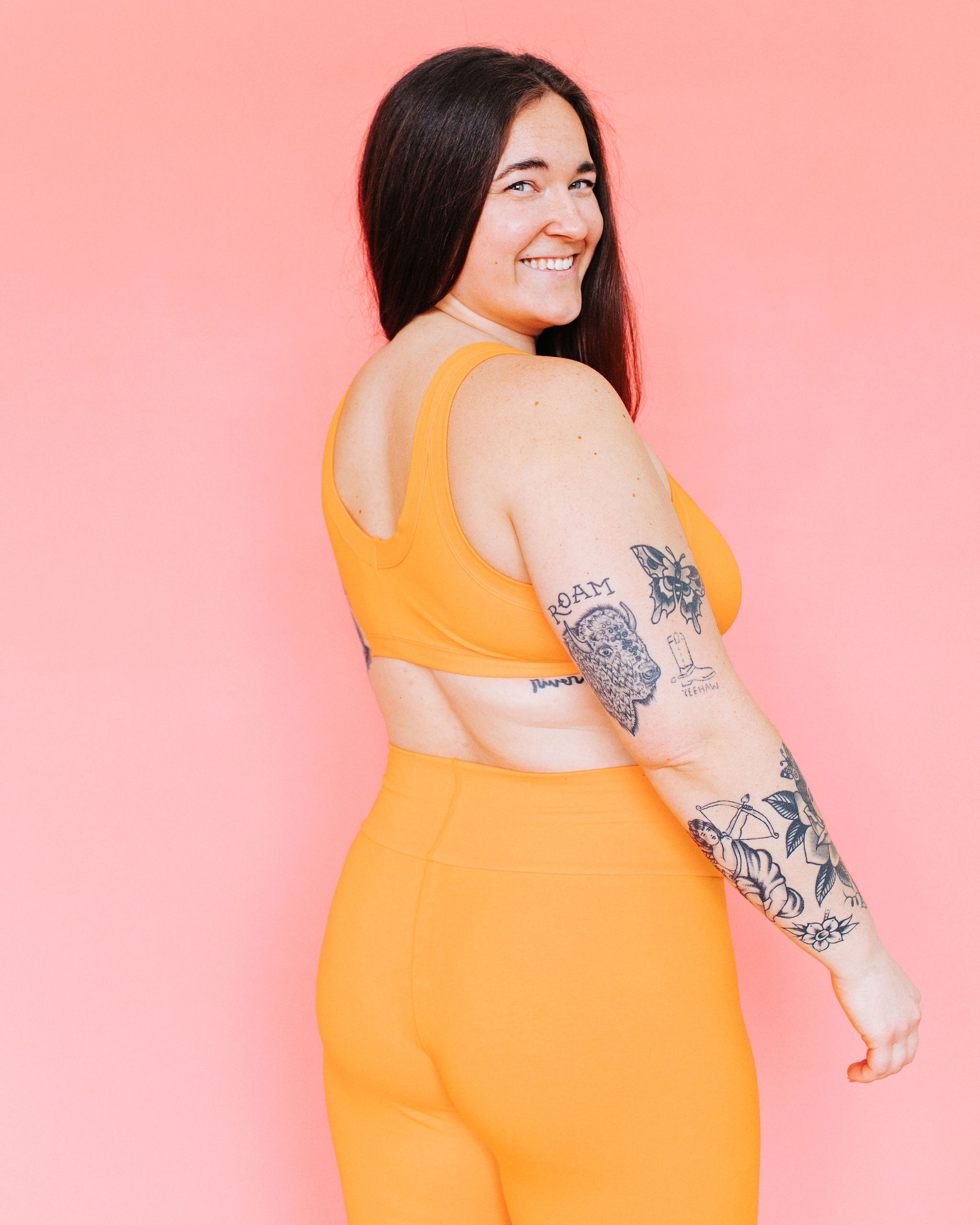 Model standing with their back to us in front of a pink background wearing a Bralette and Leggings in the orange color Oregon Sunstone.