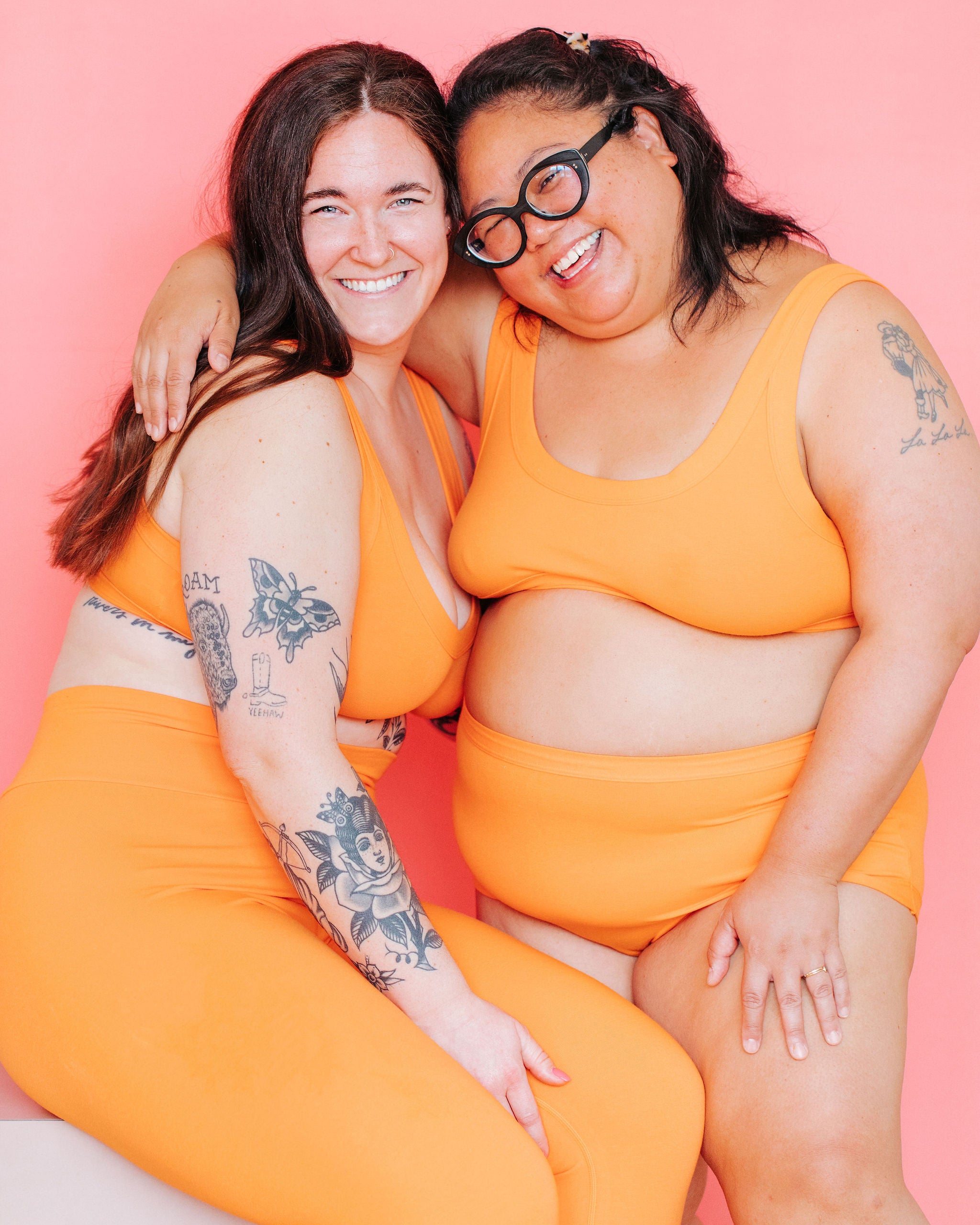 Two models sitting in front of a pink background wearing various styles in the orange color Oregon Sunstone.