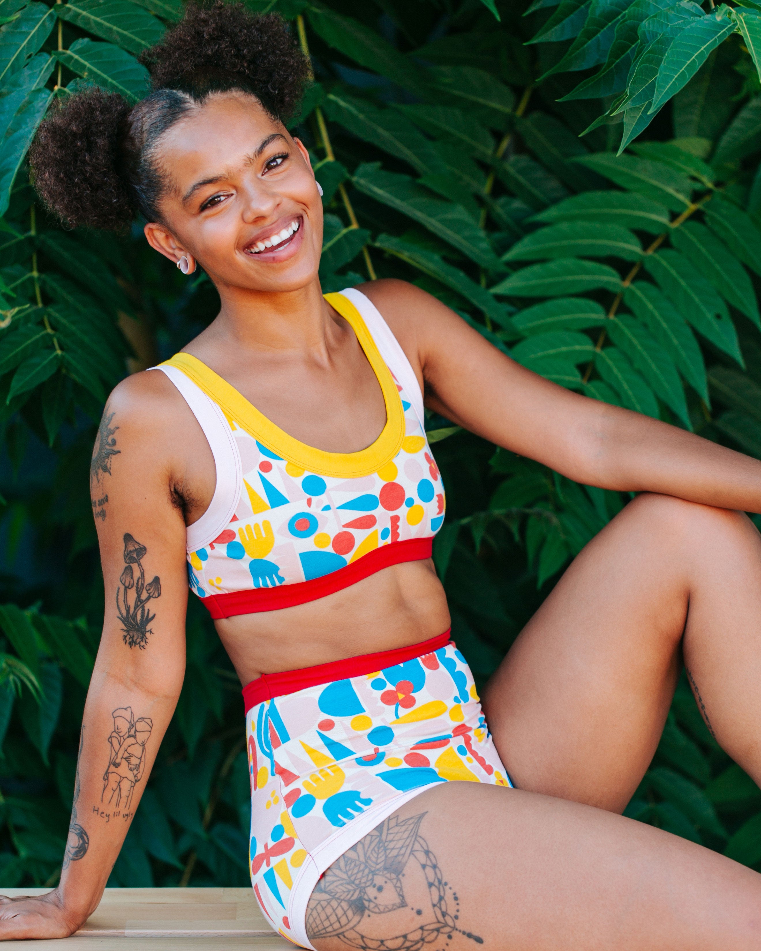 Model smiling and sitting in front of tropical plants wearing Bralette and Sky Rise style underwear in Balance by Lisa Congdon: geometric shapes in red, yellow, pink, and blue colors.