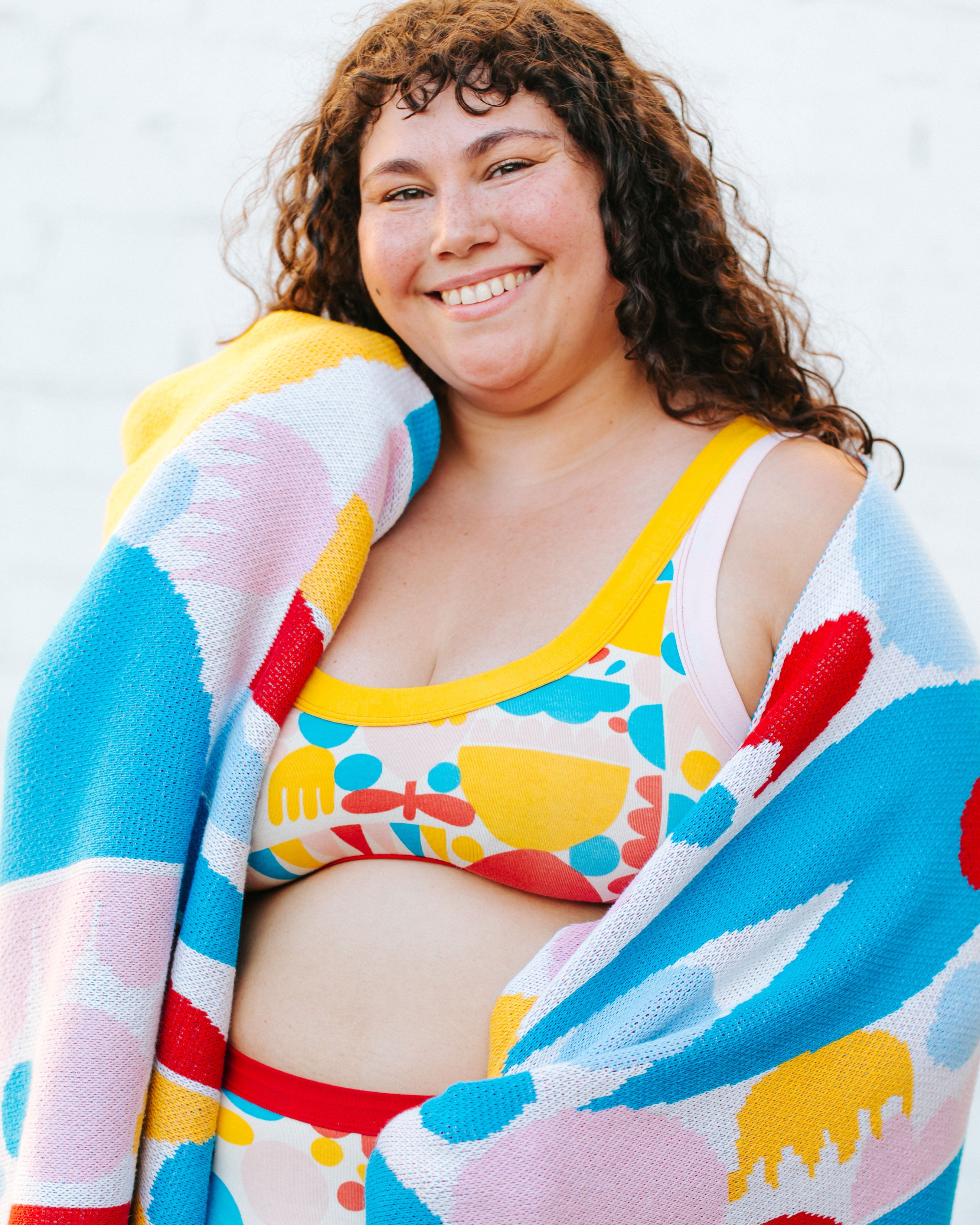 Close up of model happy wearing Bralette in Balance by Lisa Congdon: geometric shapes in red, yellow, pink, and blue colors, with a similar blanket around her shoulders.