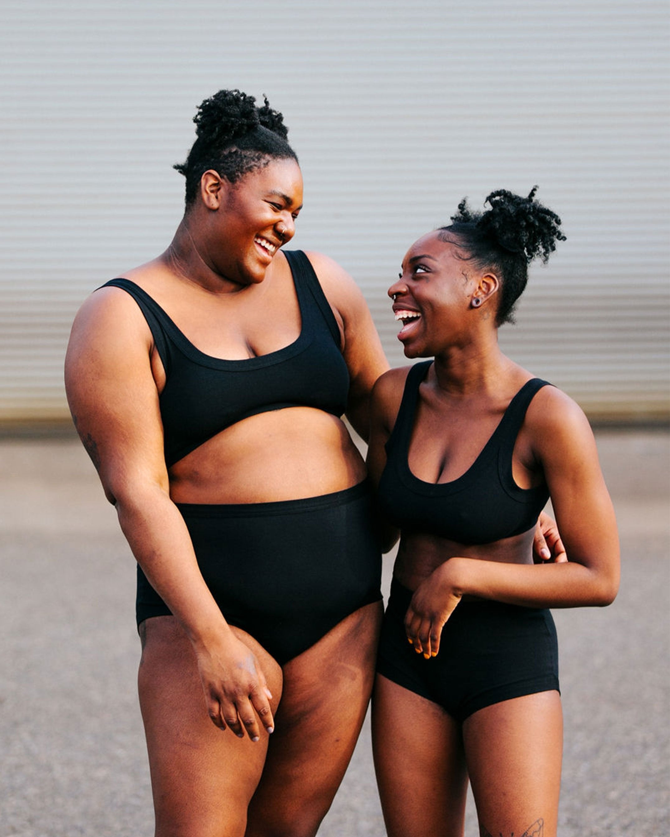 Two models looking at each other and laughing wearing Thunderpants organic cotton Bralette and Women's Original style underwear in plain black.