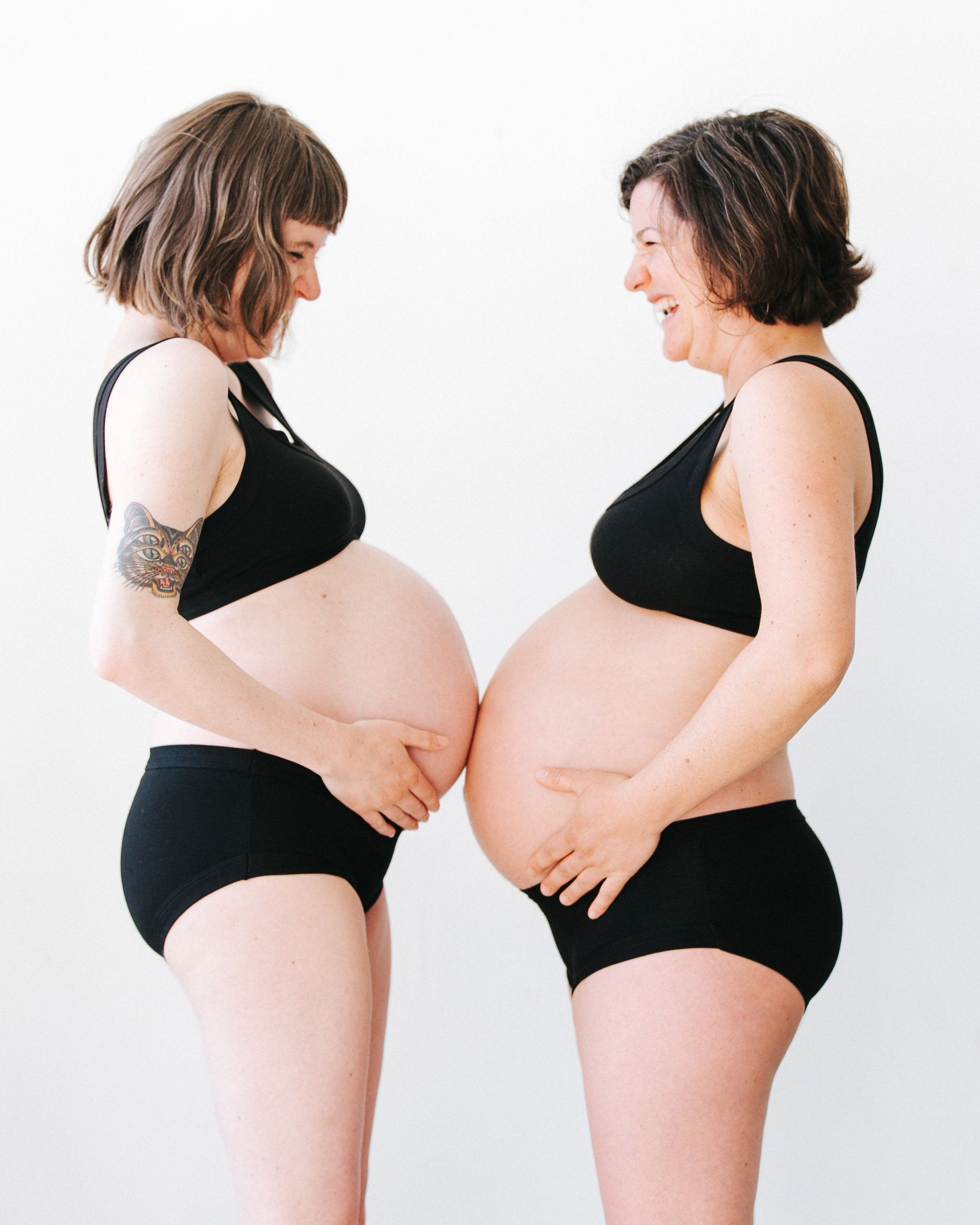 Two pregnant models standing face-to-face with their bellies touching, laughing, wearing Thunderpants organic cotton Hipster style underwear and Bralettes in plain black.