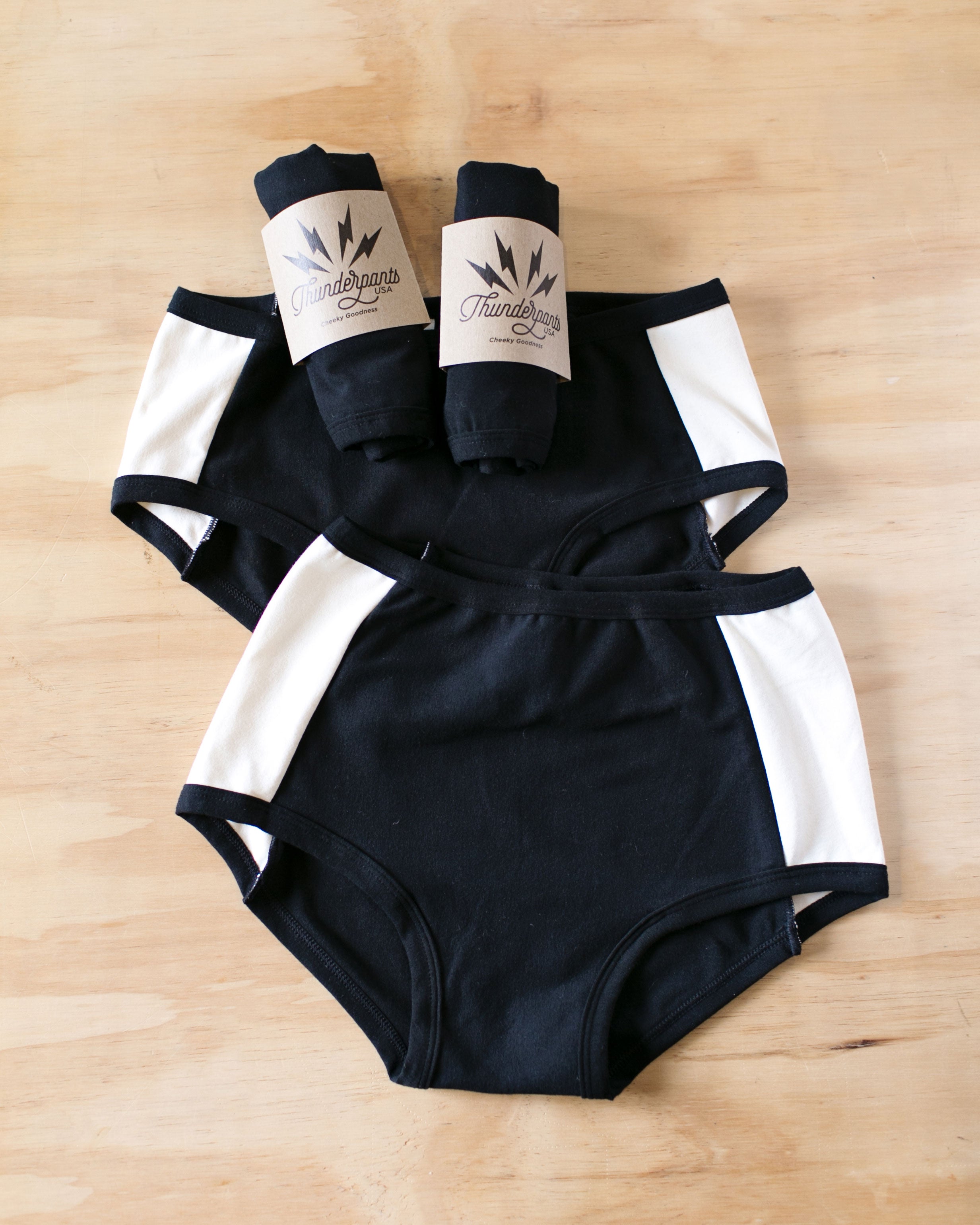 Flat lay on wood of our Cookies and Cream Panel Pants in the Hipster style, Original style, and packaged underwear: black panel in the center and vanilla panels on both sides with black binding.