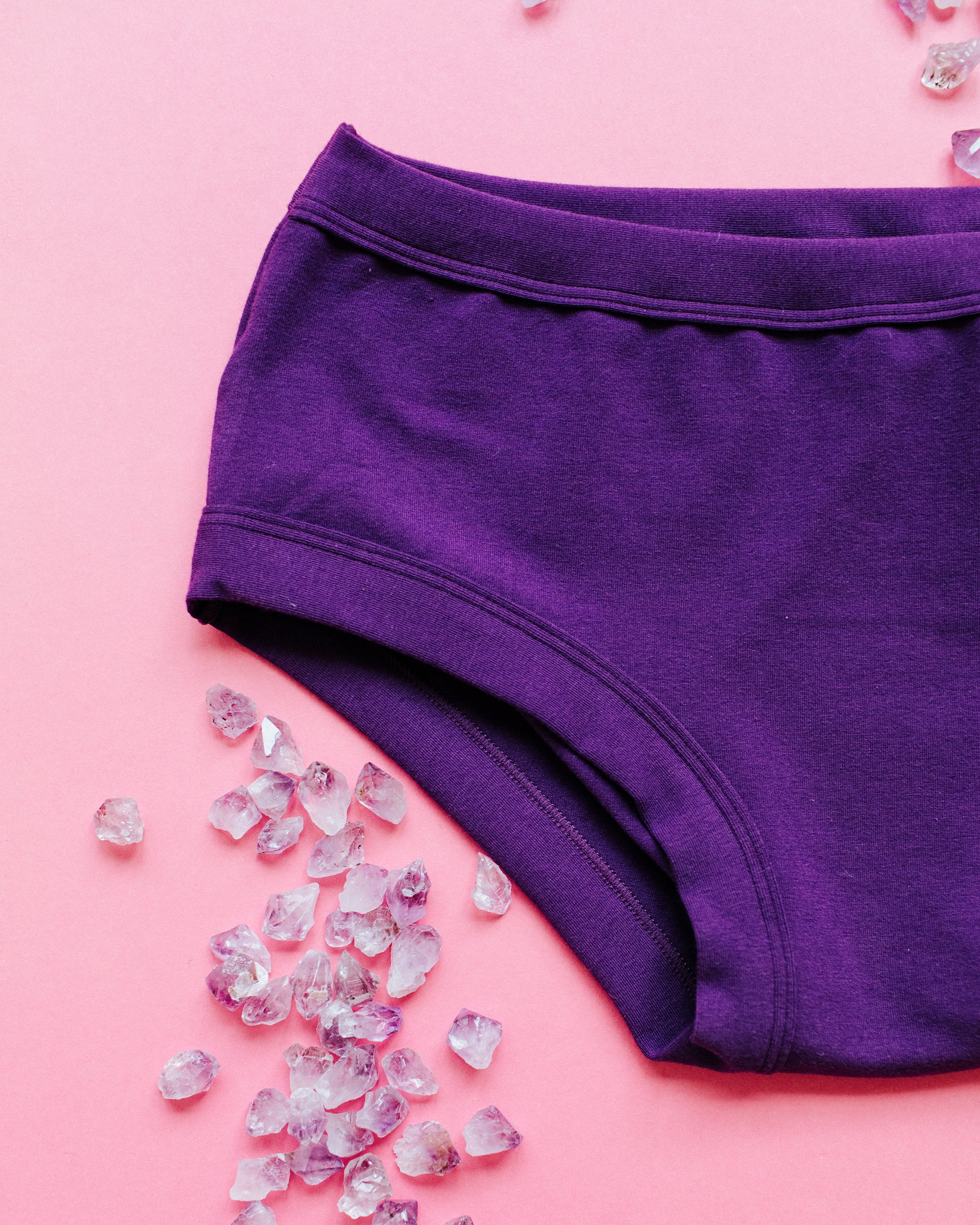 Close up flat lay of purple Deep Amethyst Hipster style underwear on a pink surface with small amethyst stones surrounding it.