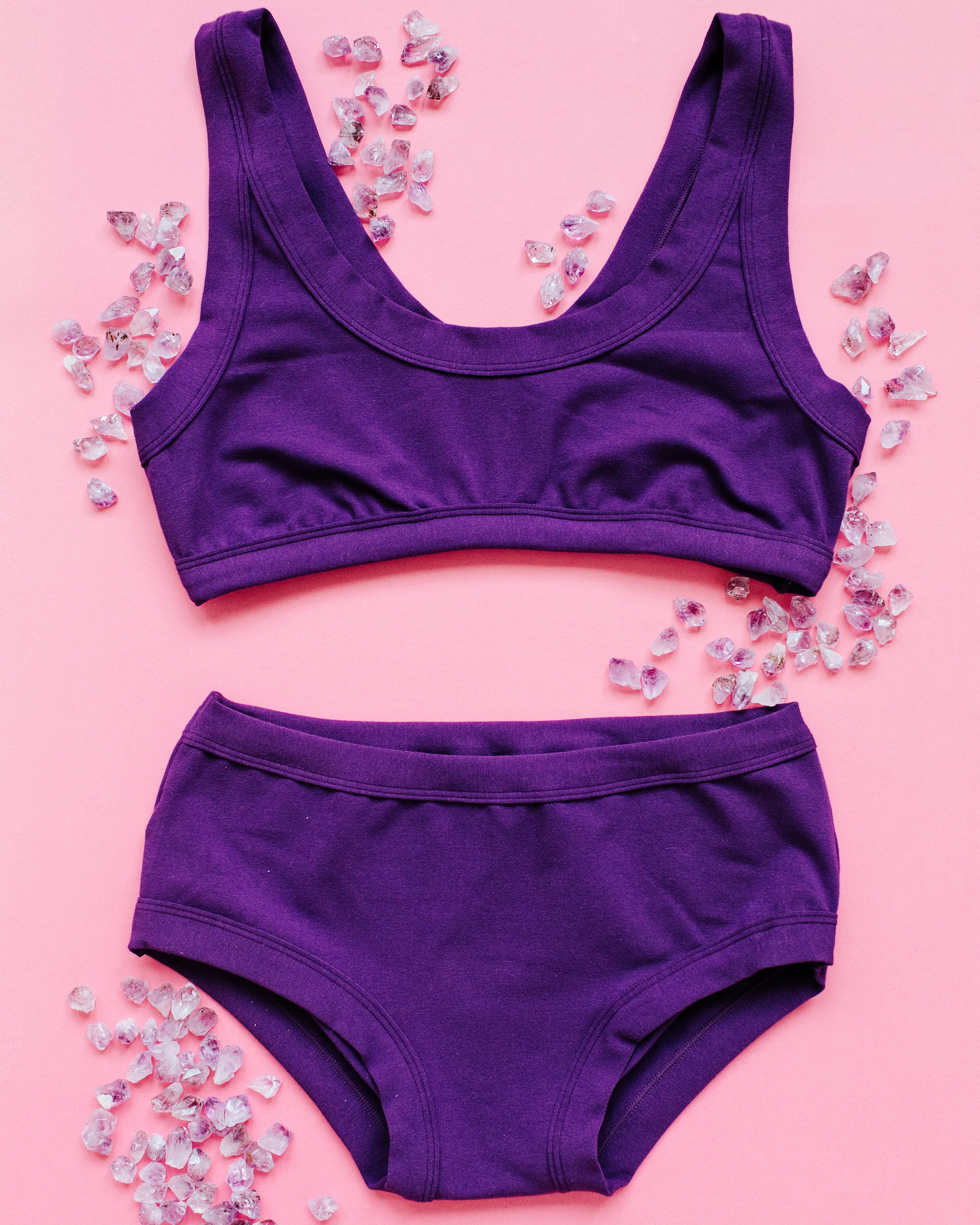 Flat lay of purple Deep Amethyst Hipster style underwear and Bralette on a pink surface with small amethyst stones surrounding.