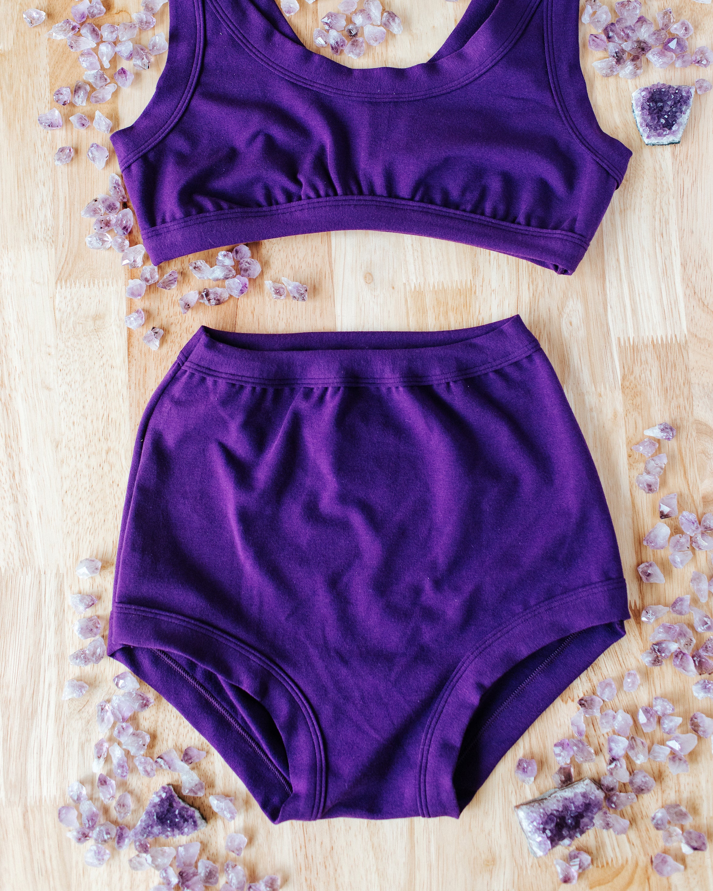 Flat lay of purple Deep Amethyst Sky Rise style underwear on a wood surface with large and small amethyst stones surrounding it.