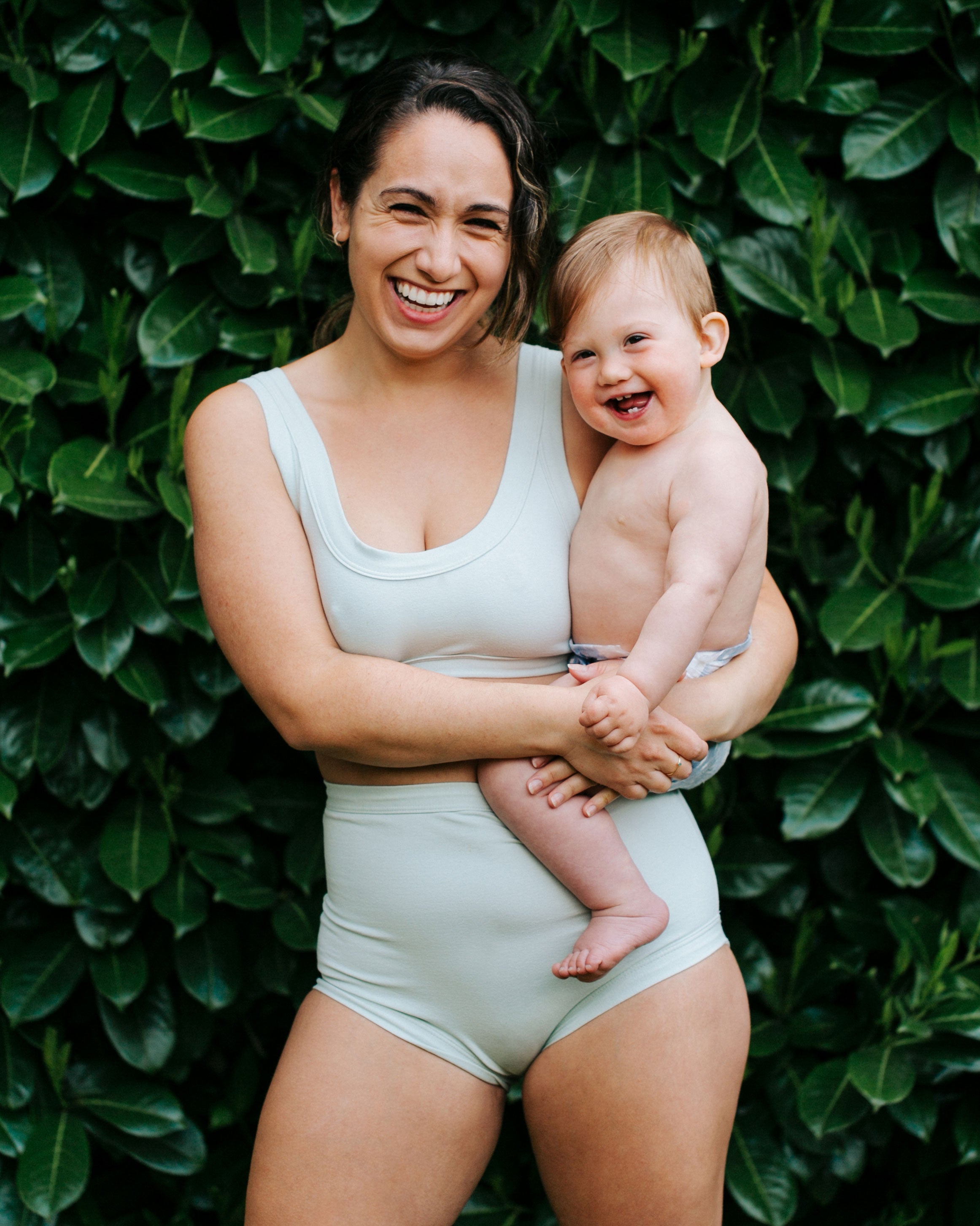 Model laughing while holding her smiling baby in front of green leaves wearing a set of Dried Sage Bralette and Sky Rise style underwear.