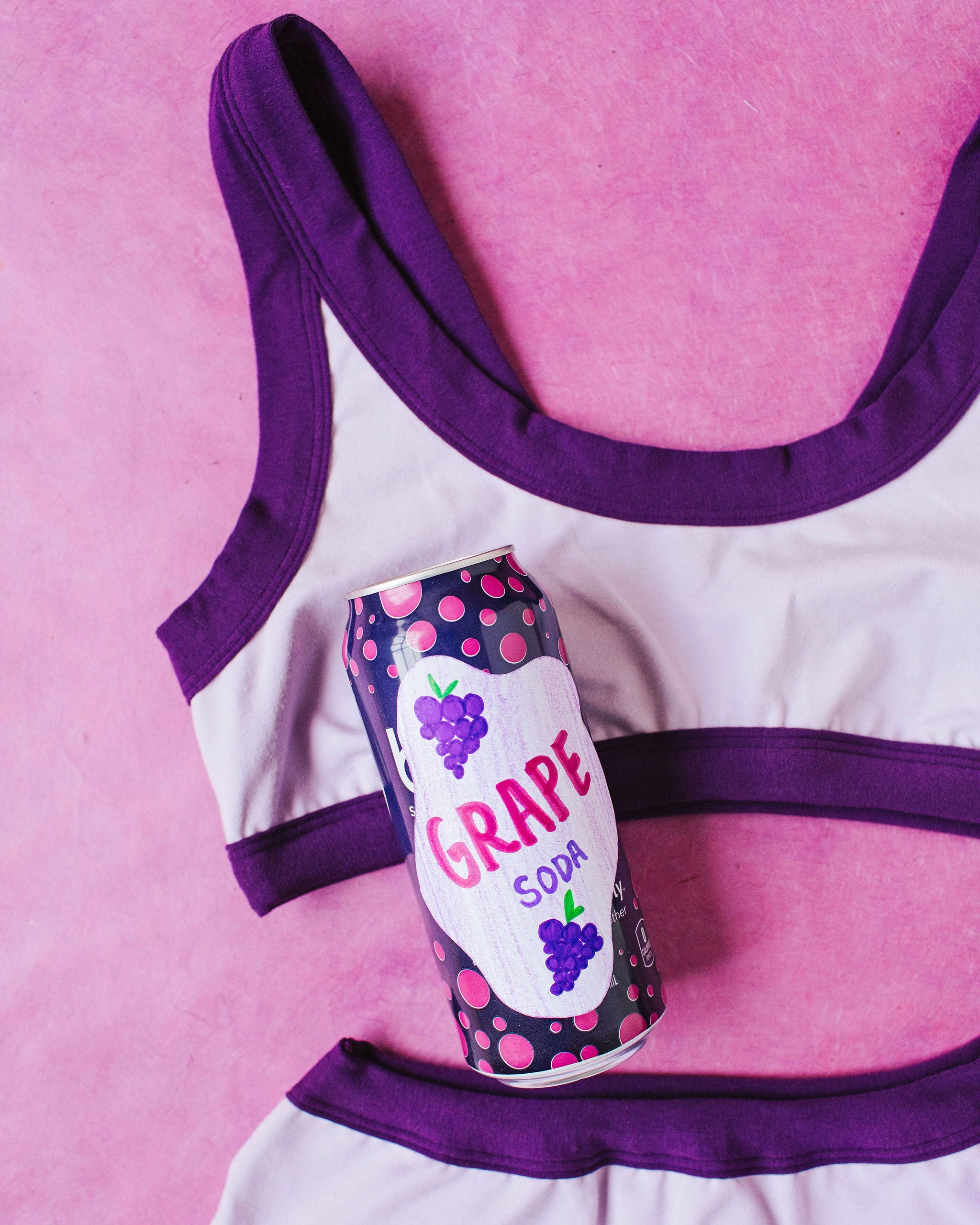 Flat lay with a grape soda can on a purple surface of Grape Soda Bralette: Lavender color with dark purple binding.