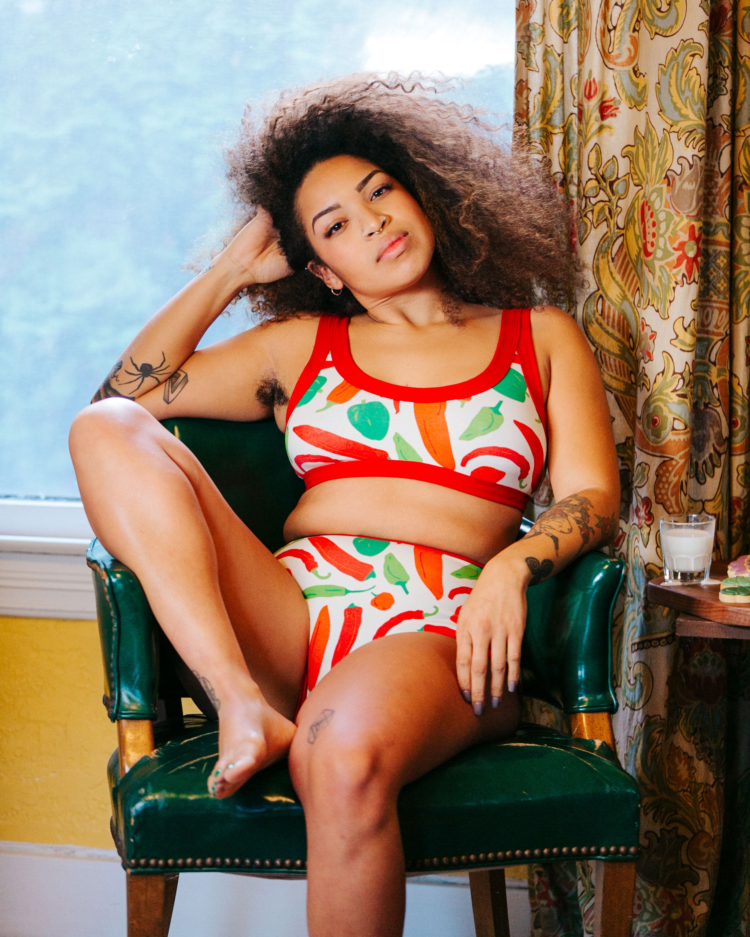 Model sitting in a chair at home wearing Thunderpants organic cotton Bralette and Sky Rise style underwear in our Hot Pants print: various green, orange, and red peppers printed on Vanilla with red binding.