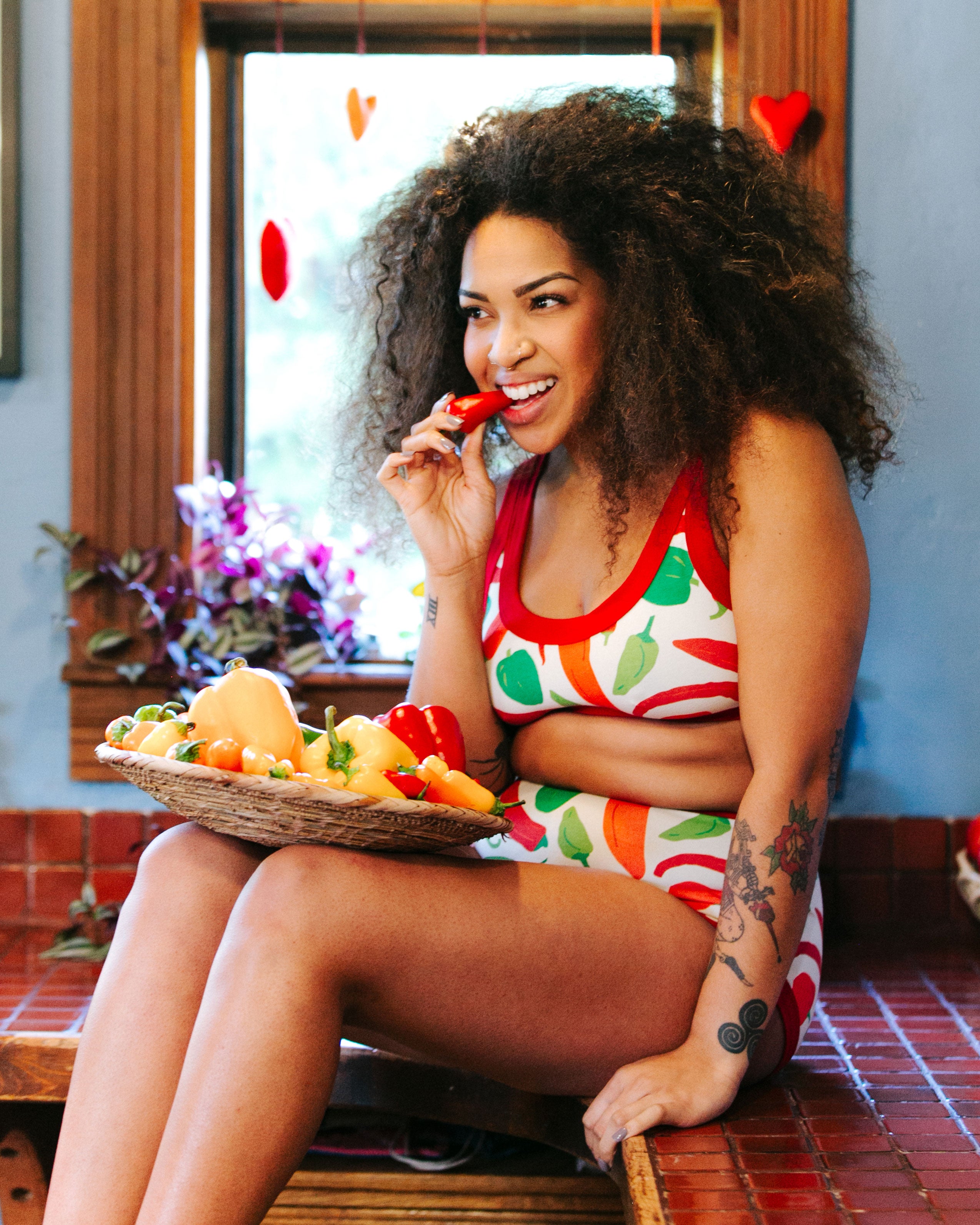 Model sitting on a kitchen counter eating a pepper wearing Thunderpants organic cotton Bralette and Sky Rise style underwear in our Hot Pants print: various green, orange, and red peppers printed on Vanilla with red binding.
