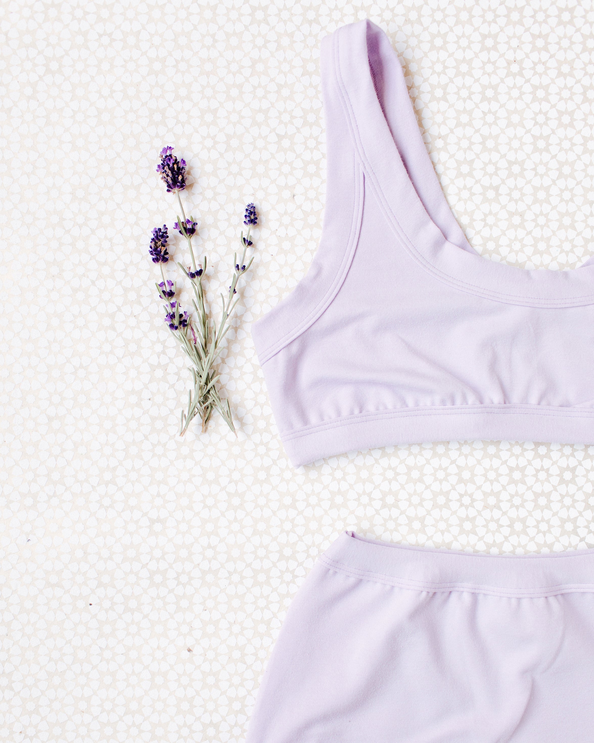 Close up flat lay of Light Lavender Bralette on a white patterned surface with small lavender sprigs.