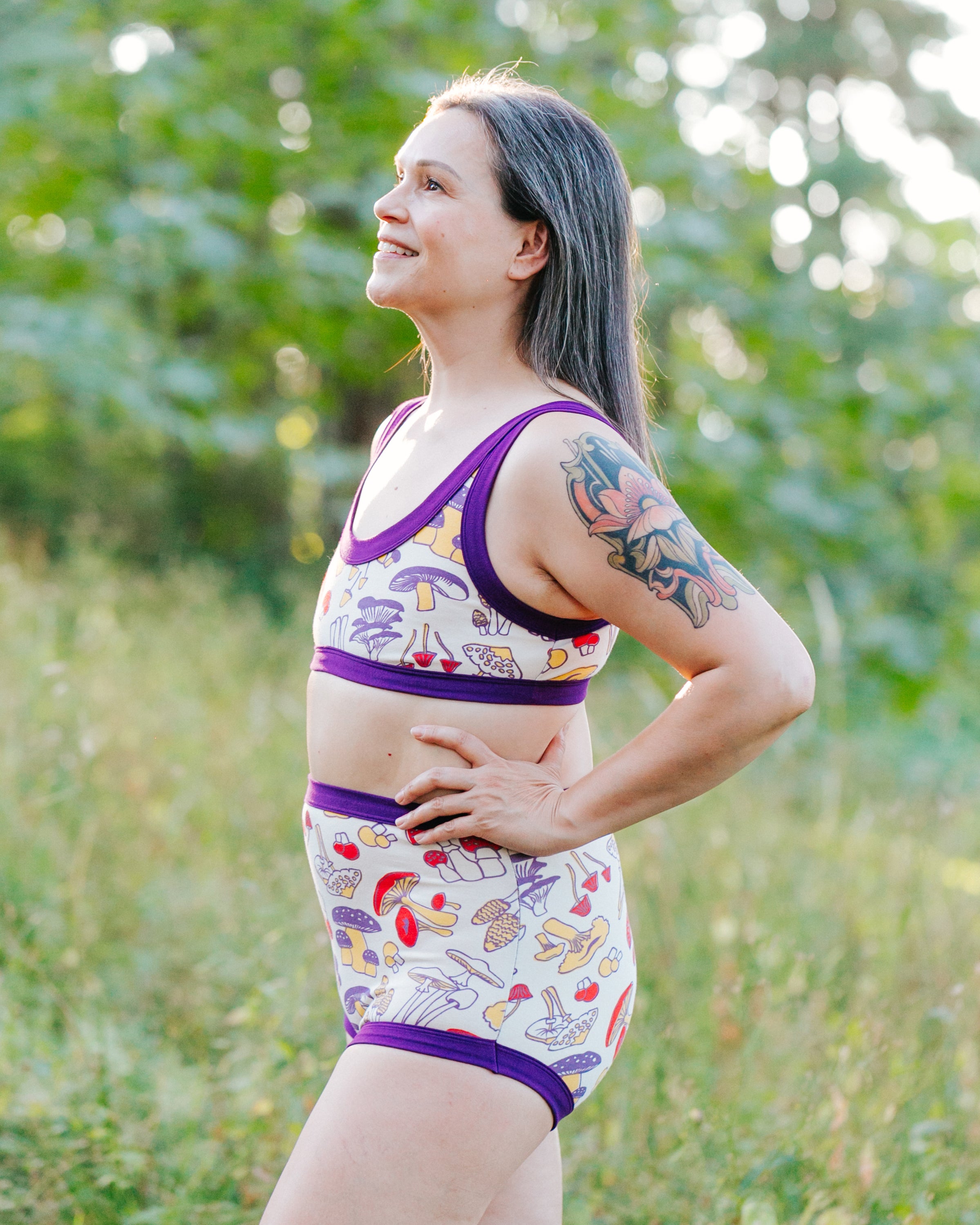 Model in the forest wearing a set of Bralette and Sky Rise style underwear in Mushroom Magic print: different kinds of mushrooms in red, yellow, and purple colors with dark purple binding.