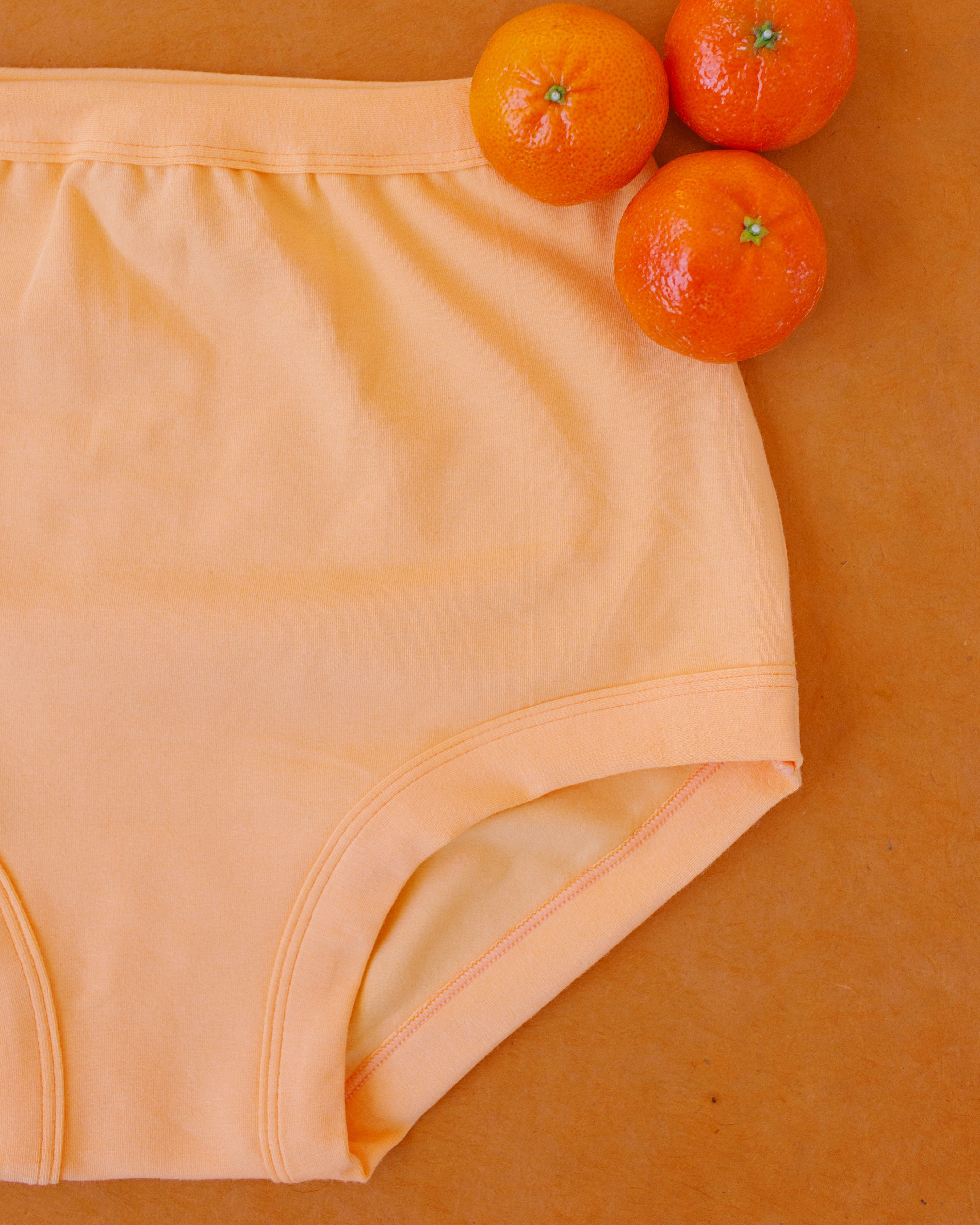 Flat lay of Orange Sherbet Sky Rise style underwear on an orange surface with oranges.