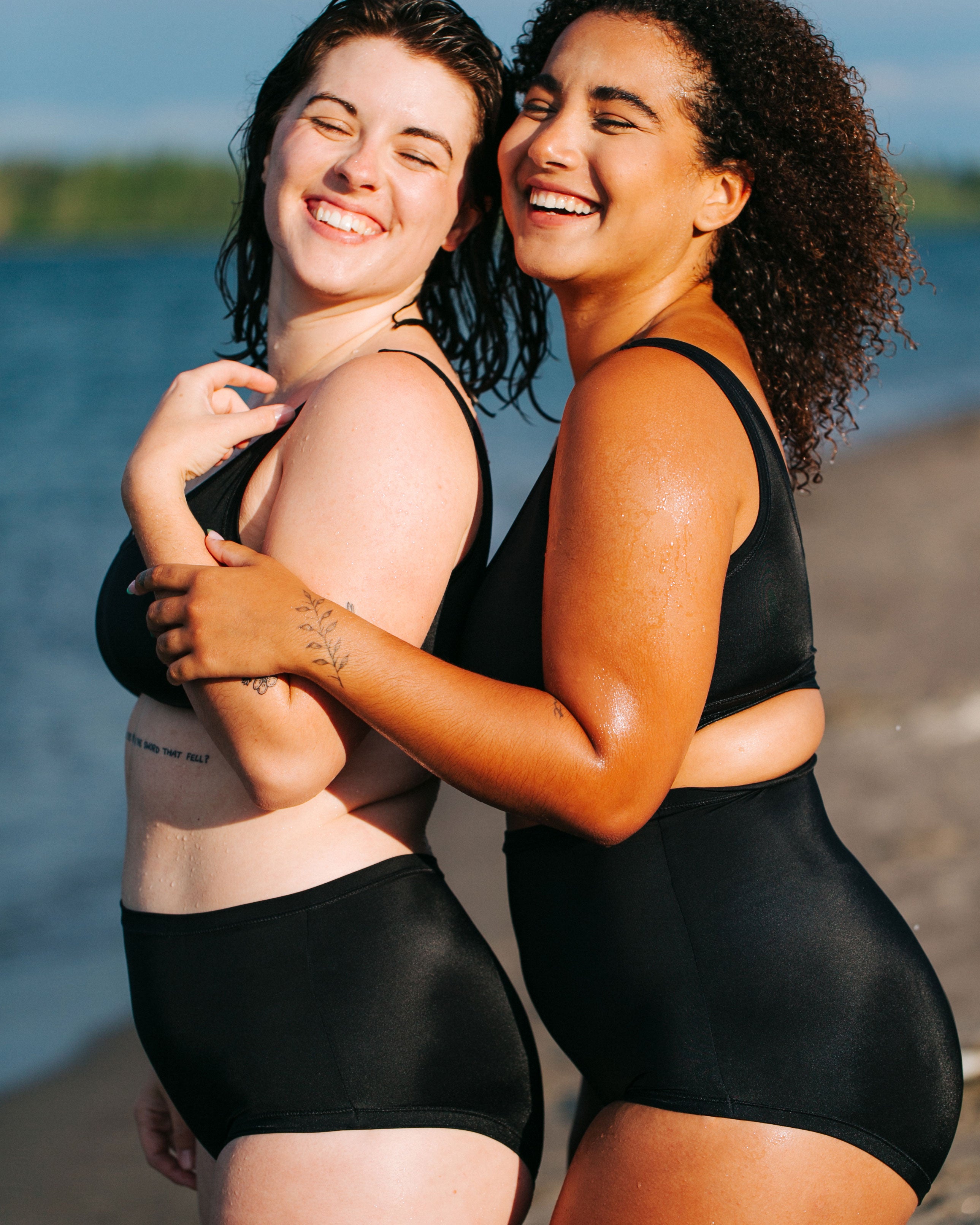 Two models from the side laughing together on the beach wearing Thunderpants recycled nylon Swimwear Original style Bottoms and Sky Rise style , and Swimwear Top in Plain Black.