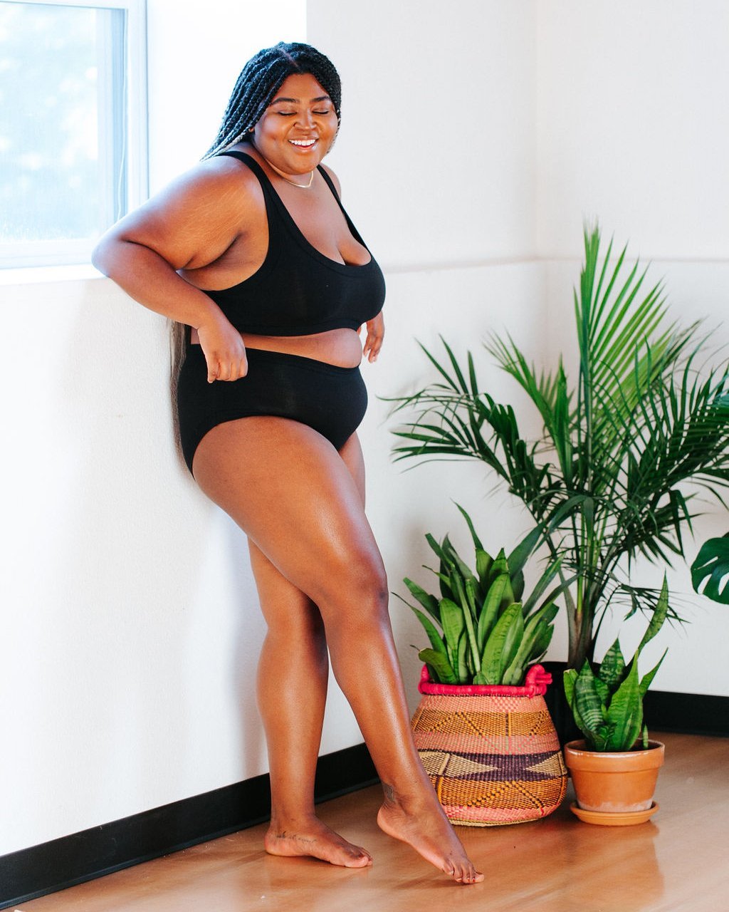 Smiling plus-sized model wearing Thunderpants organic cotton Hipster style underwear and Bralette in plain black surrounded by plants.