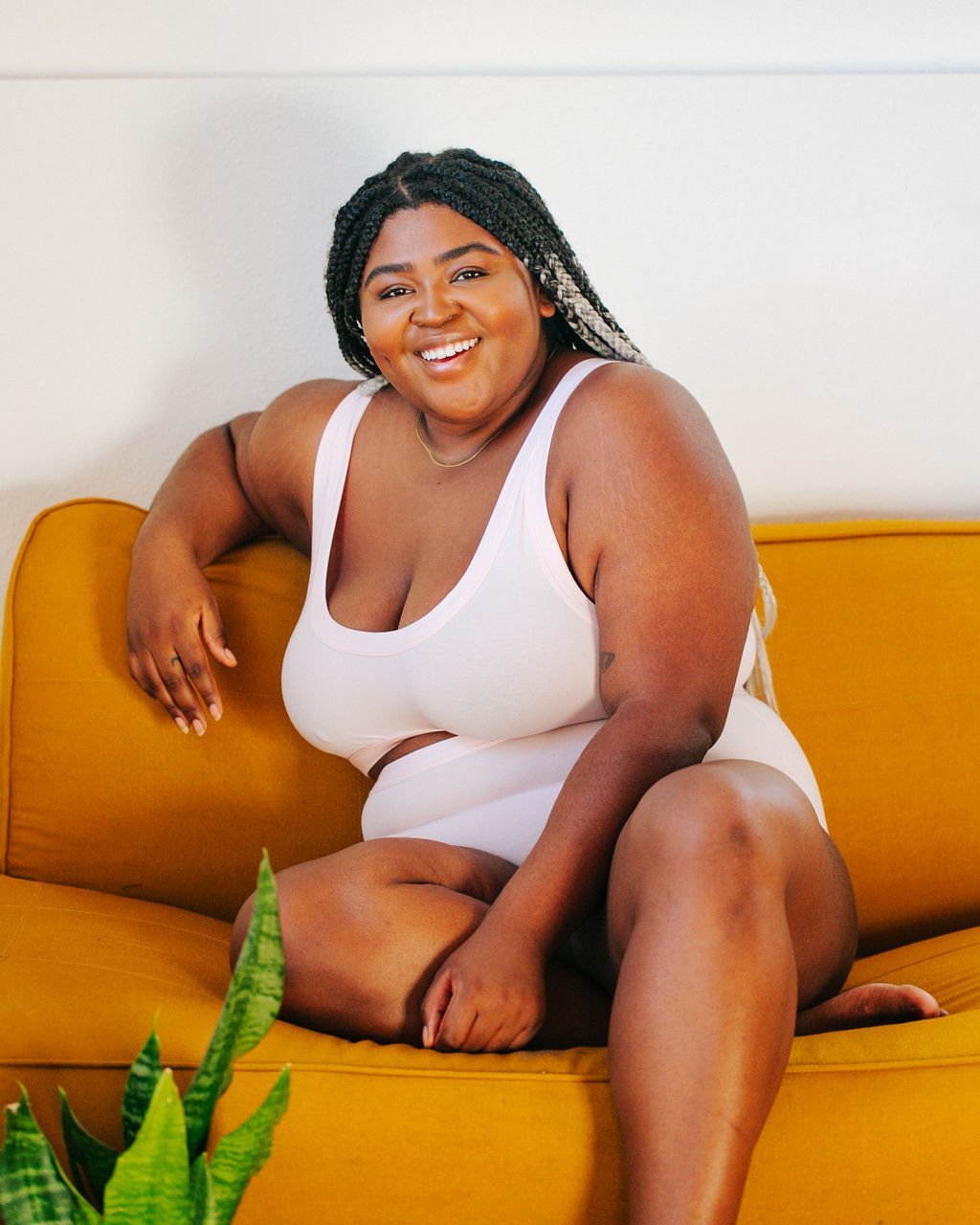Happy and beautiful plus-sized model wearing Thunderpants organic cotton Bralette and Women's Original style underwear in plain pink sitting on a bright yellow couch.