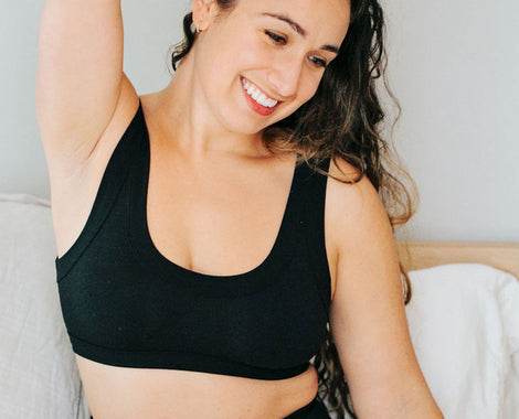 Model smiling while sitting on a bed wearing Thunderpants Bralette in Plain Black.