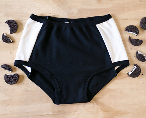 Flat lay on a wood surface and oreos around of Original style Panel Pants underwear in Cookies and Cream: black front and back with vanilla sides.