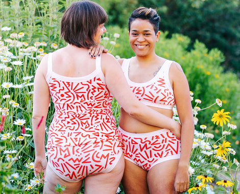 Two models, one facing us and one facing away, wearing Thunderpants Organic Cotton Original and Hipster Underwear, Bralette and Camisole in our Energy Vibes print: big pink squiggly lines over Vanilla.
