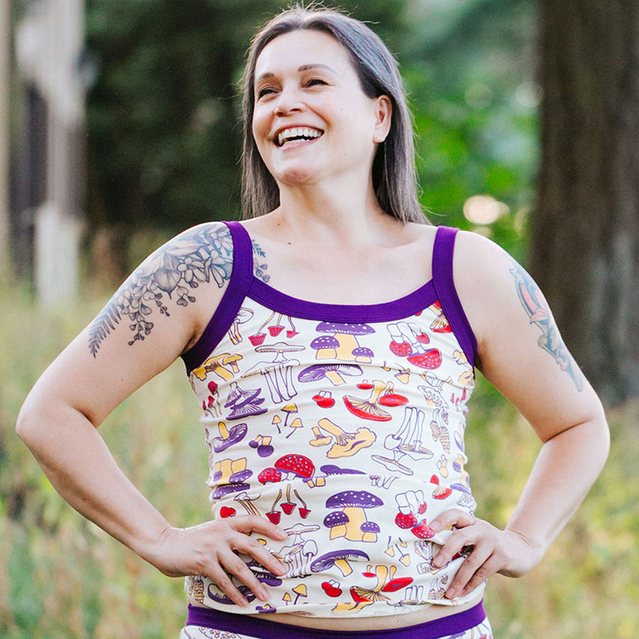 Model smiling in the forest wearing a set of Camisole and Hipster style underwear in Mushroom Magic print: different kinds of mushrooms in red, yellow, and purple colors with dark purple binding.