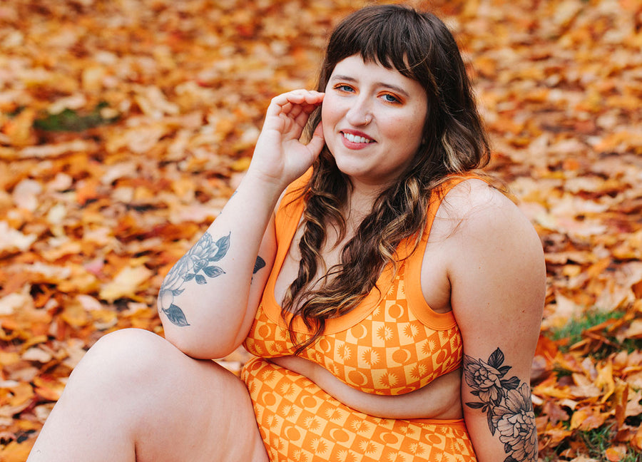 Model sitting in fall orange leaves wearing Thunderpants Sky Rise style and Bralette in Autumn Equinox print: sun and moon pattern in two oranges.