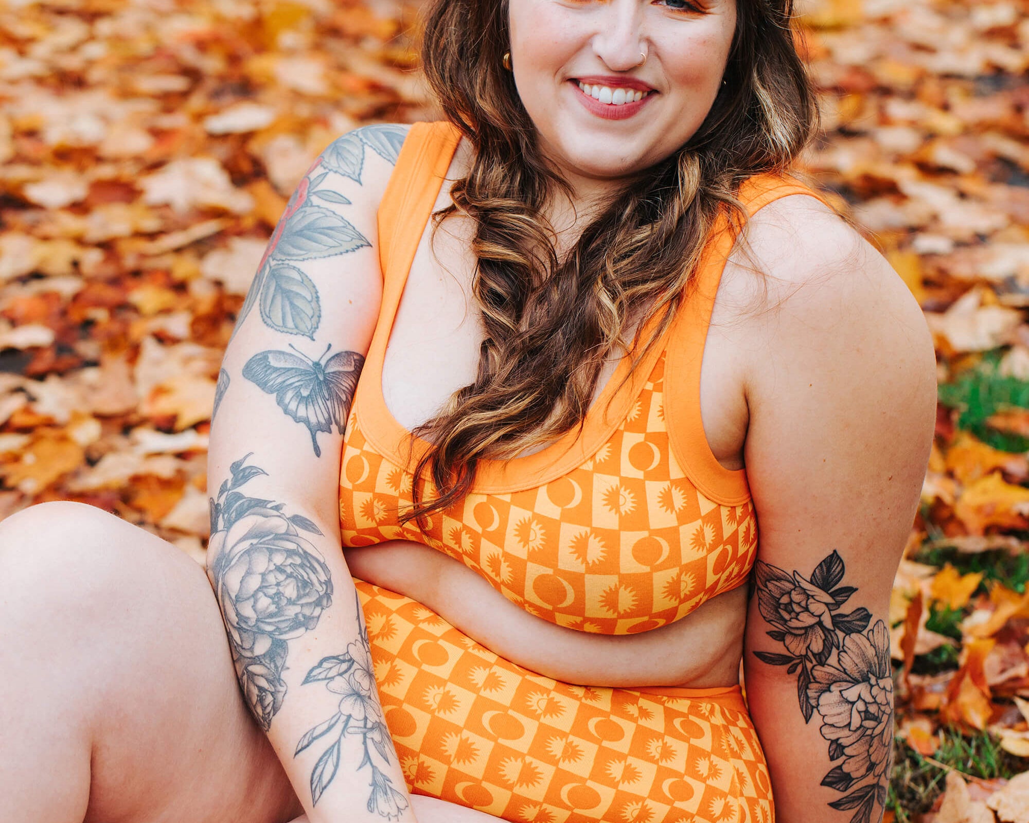 Model sitting in a bunch of fall leaves wearing a set of Thunderpants Bralette and Sky Rise style underwear in Autumn Equinox print. 