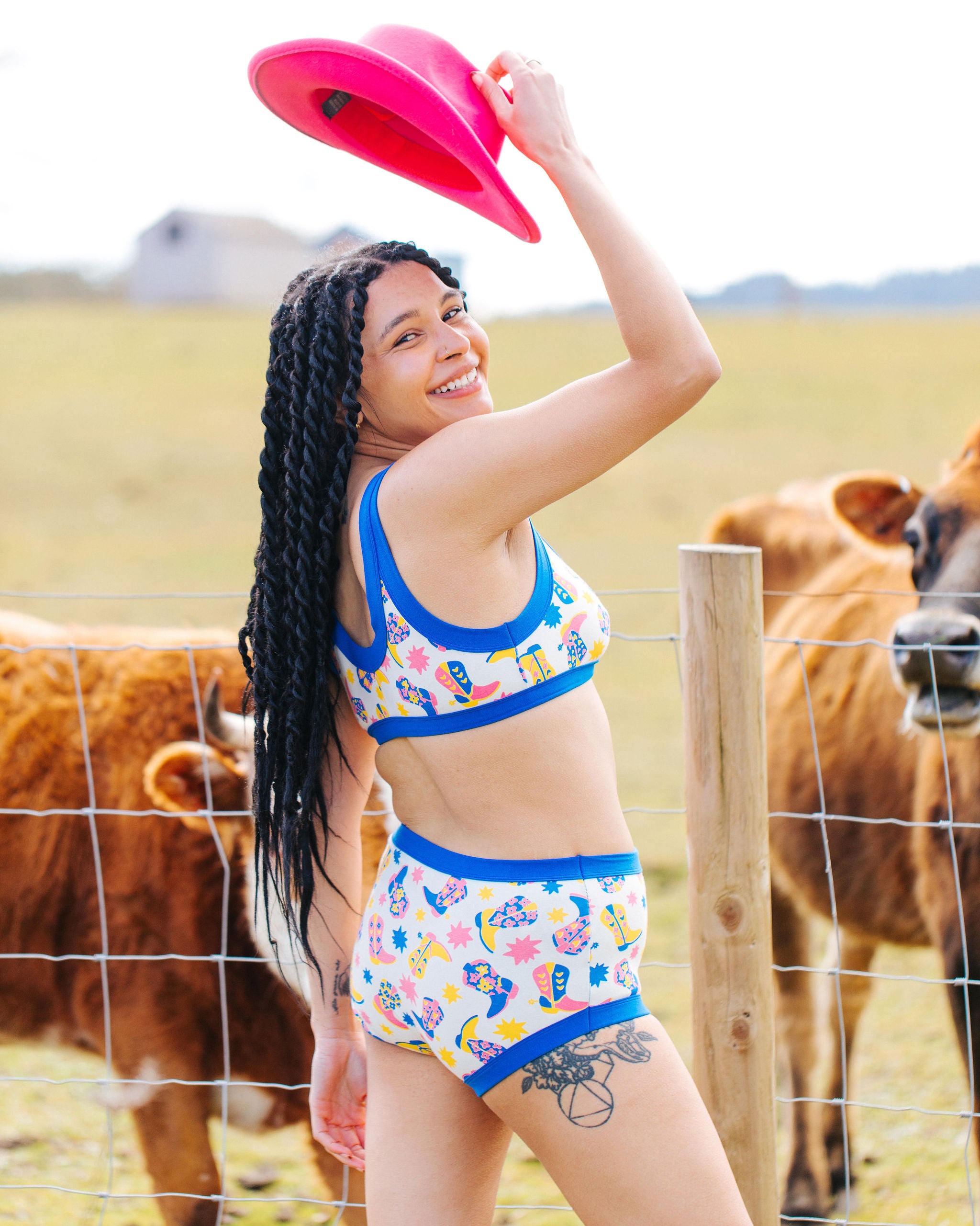 Model smiling with cows wearing Thunderpants Bralette and Original style underwear in Boot Scootin' - fun boots in pink, yellow, and blue.