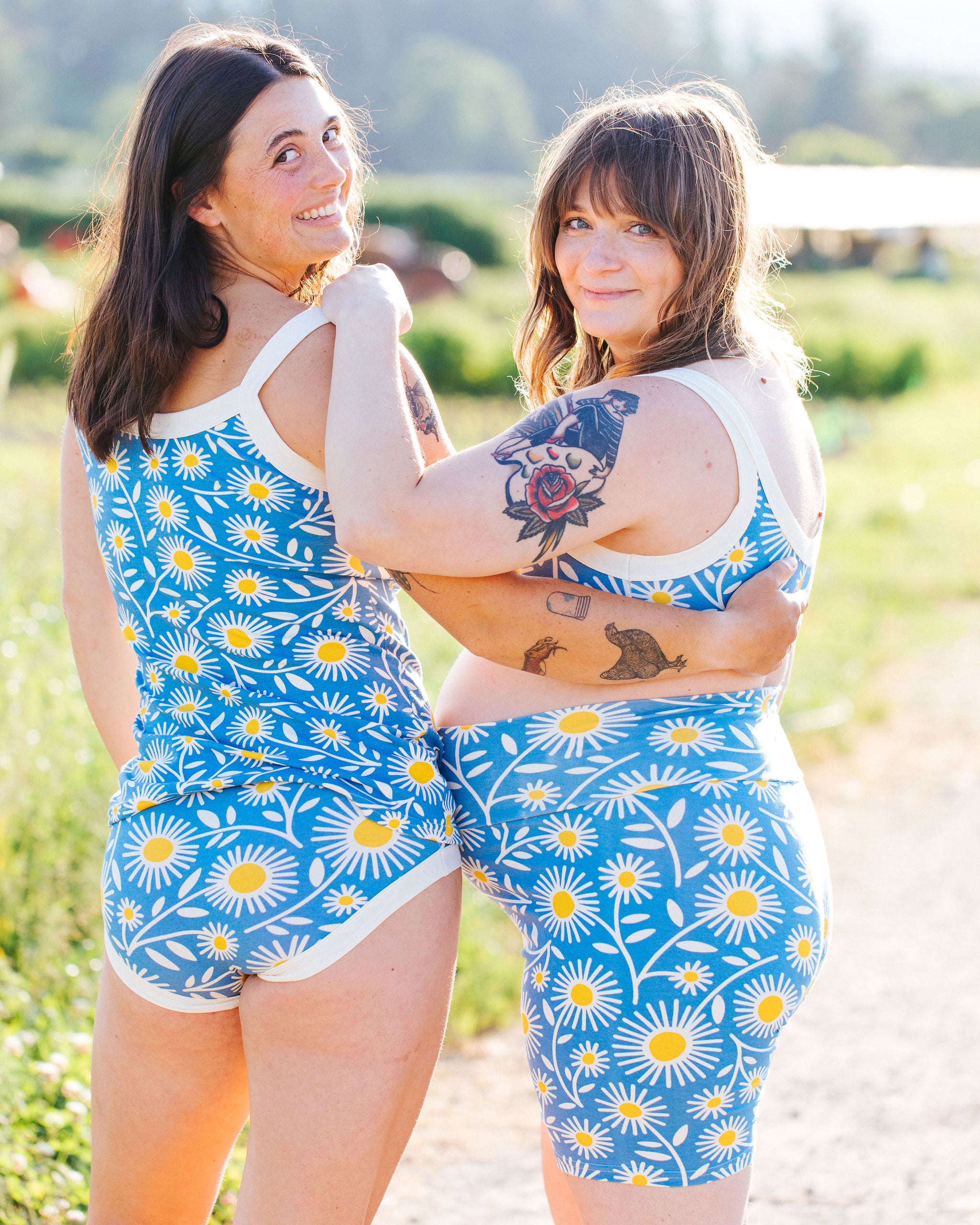 Two models smiling in a field wearing Thunderpants Bike Shorts in Daisy Days print: blue with white and yellow daisies.