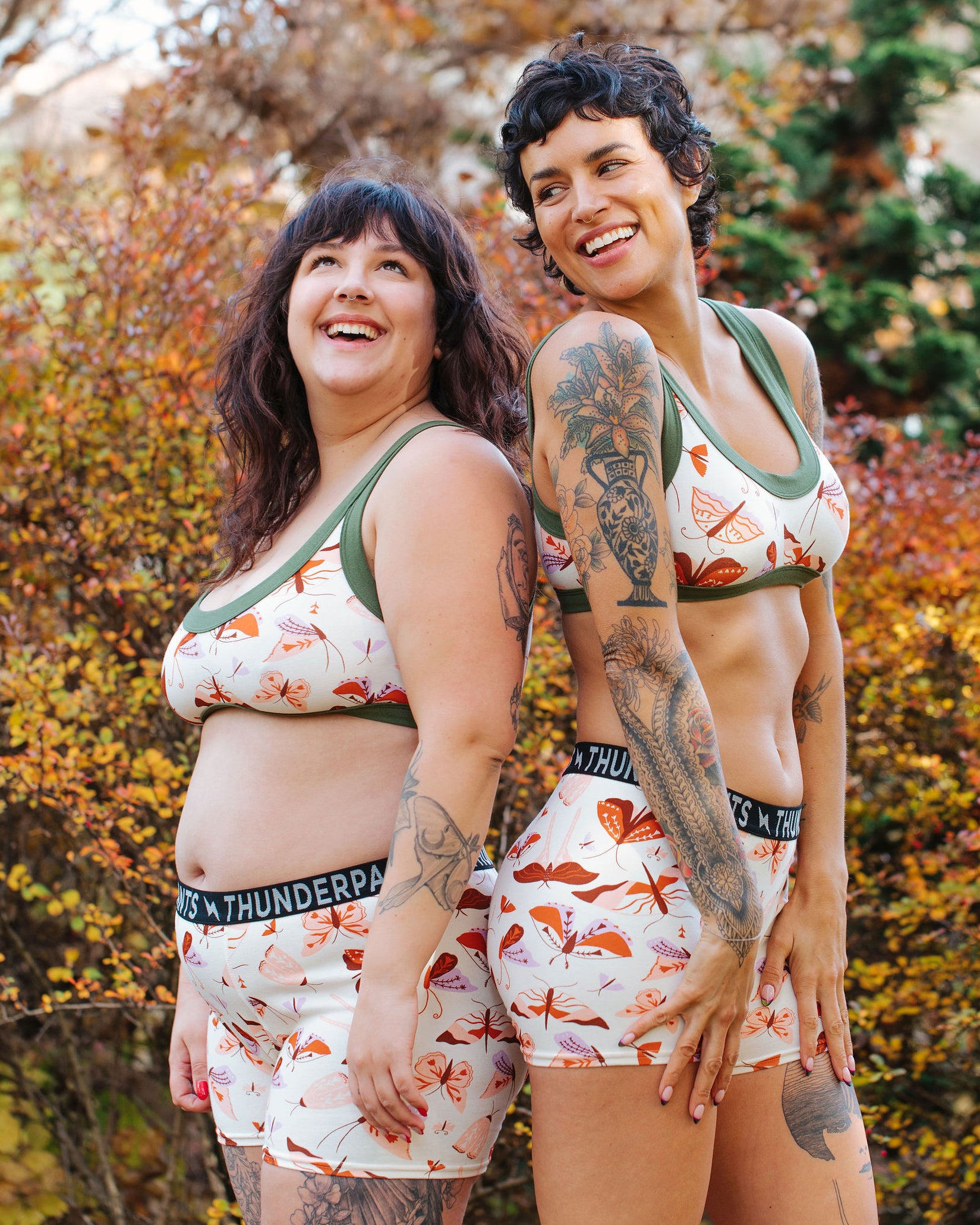 Models wearing Thunderpants Bralette and Boxer Briefs in Field Study: a moth print in red, orange, pink, and purple bound in green.
