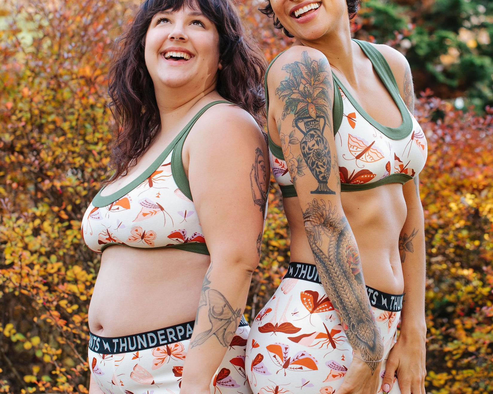 Models wearing Thunderpants Bralette and Boxer Briefs in Field Study: a moth print in red, orange, pink, and purple bound in green.