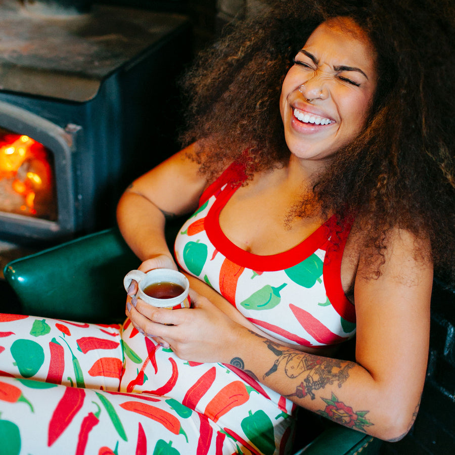 Model holding tea in a chair by the fire wearing Thunderpants Organic Cotton Leggings and Bralette in our Hot Pants print: natural with red, orange, and green peppers with red binding.