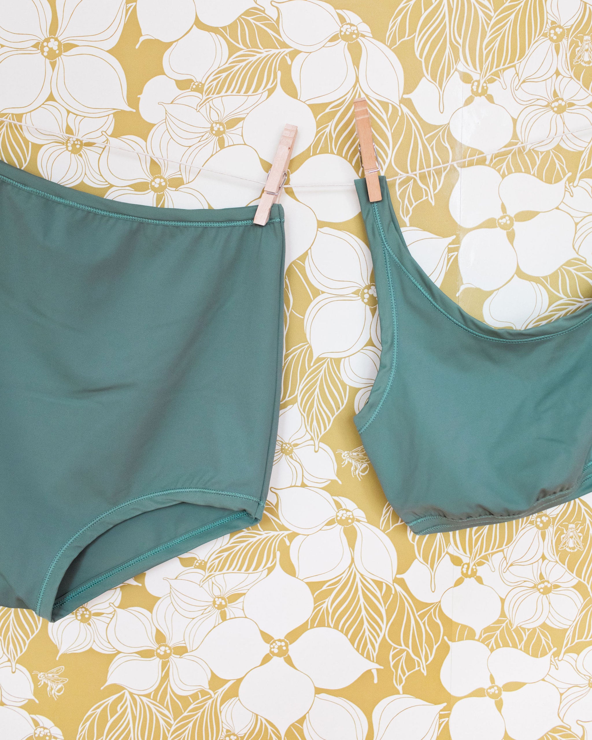 Swimwear Sky Rise Bottoms and Top in Lichen Green hanging with clothes pins on a floral wall.