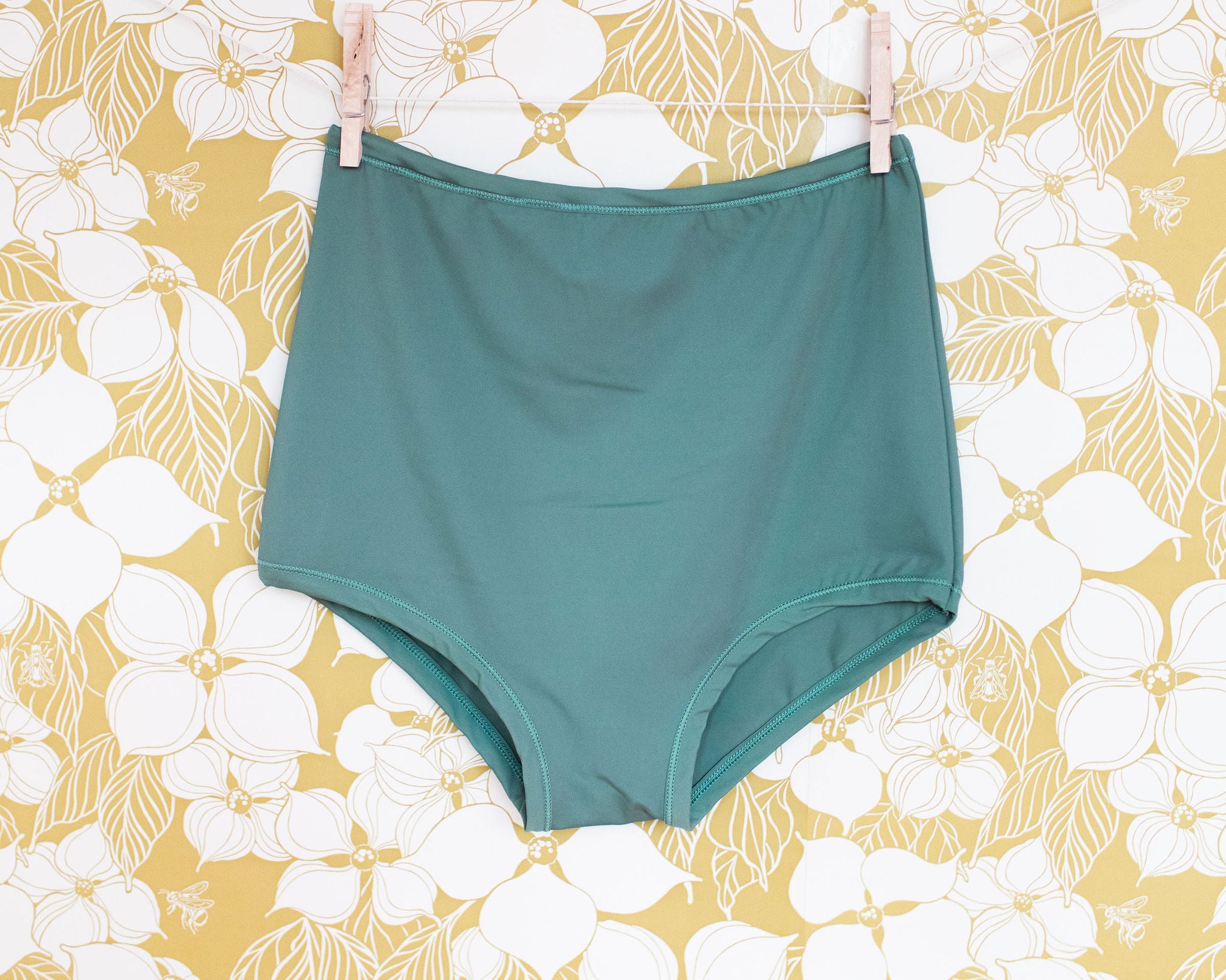 Swimwear Sky Rise Bottoms in Lichen Green hanging with clothes pins on a floral wall.