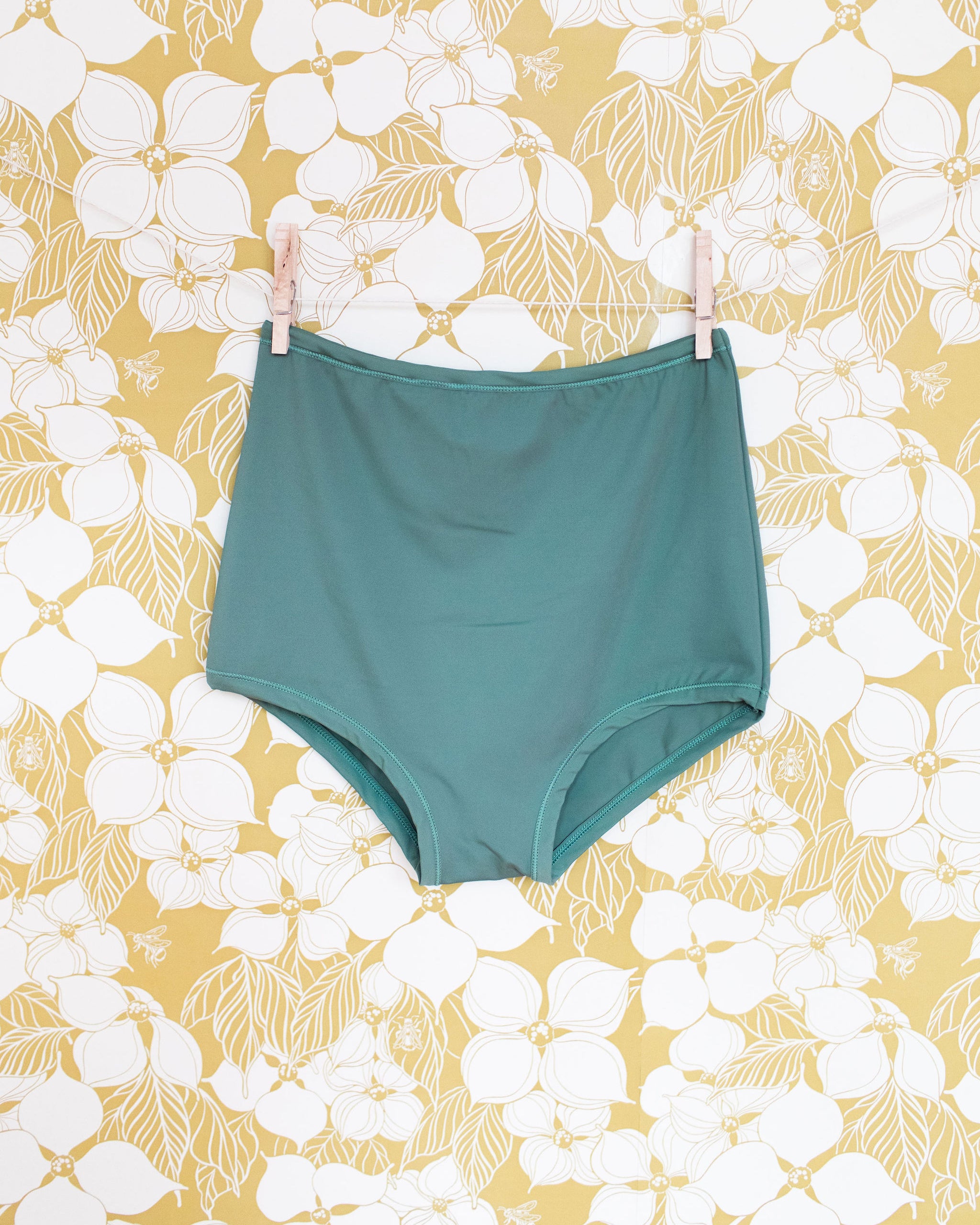 Swimwear Sky Rise Bottoms in Lichen Green hanging with clothes pins on a floral wall.