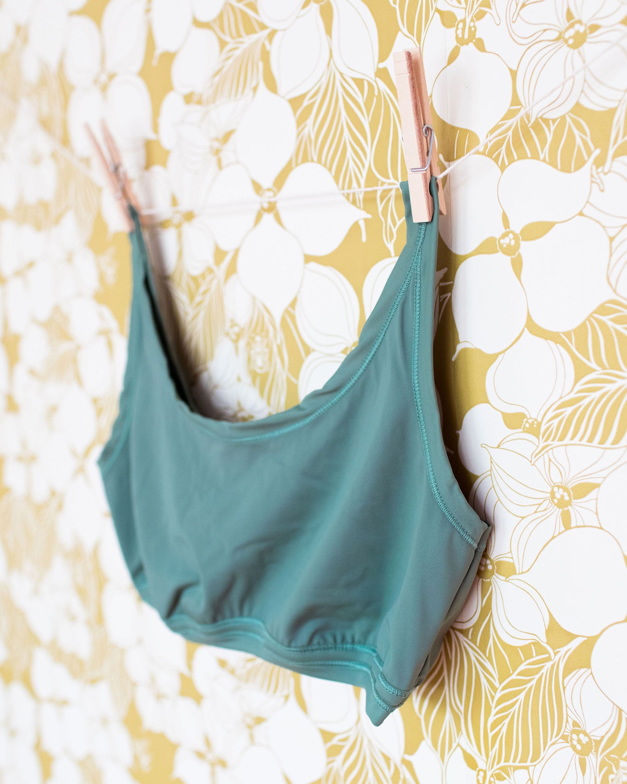 Close up of Swimwear Top in Lichen Green hanging with clothes pins on a floral wall.