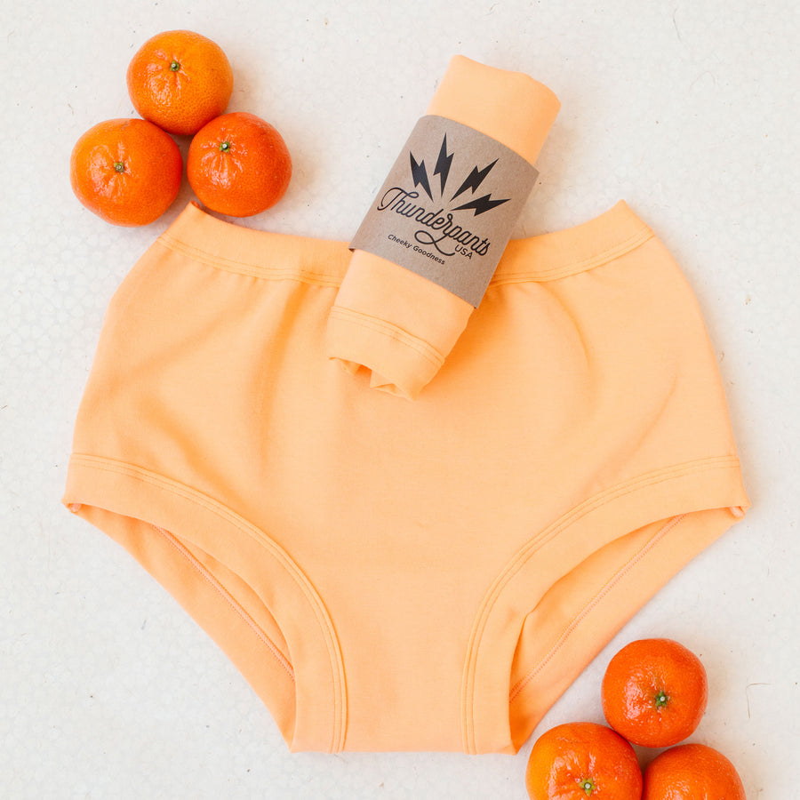 Flat lay of a packaged pair of Orange Sherbet underwear on a white and orange surface with oranges around it.