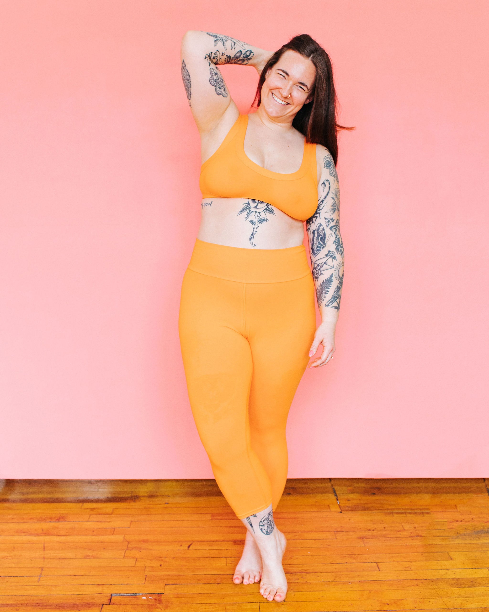 Model standing in front of a pink background wearing 3/4 Length Leggings and a Bralette in the orange color Oregon Sunstone.