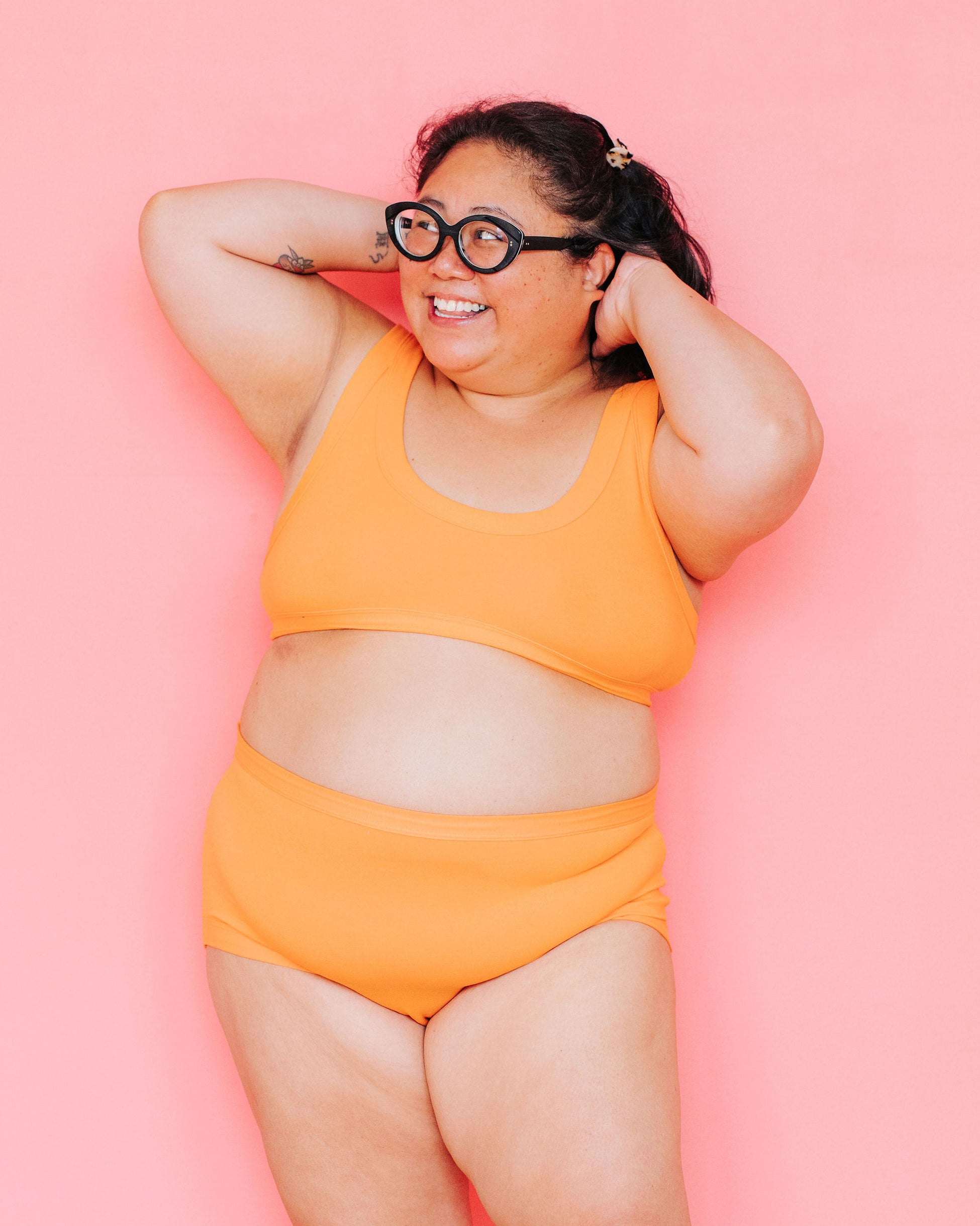 Model smiling in front of a pink background wearing the Original style underwear and a Bralette in the orange color Oregon Sunstone.
