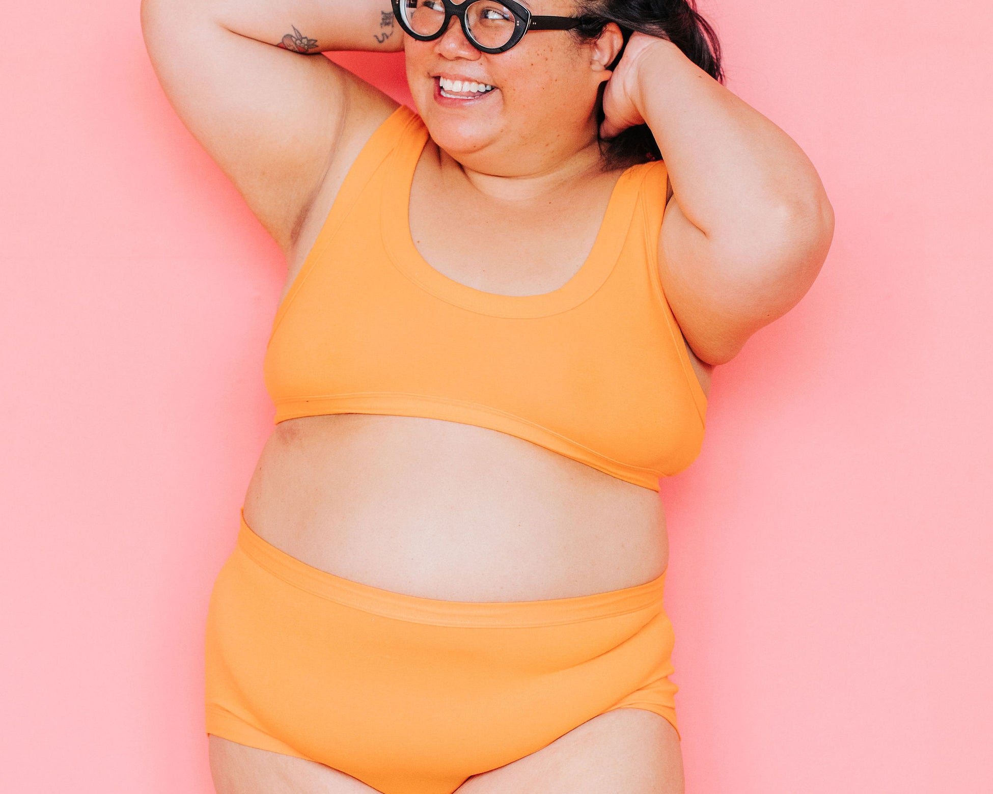 Model smiling in front of pink background wearing a Bralette and Original style underwear in the orange color Oregon Sunstone.