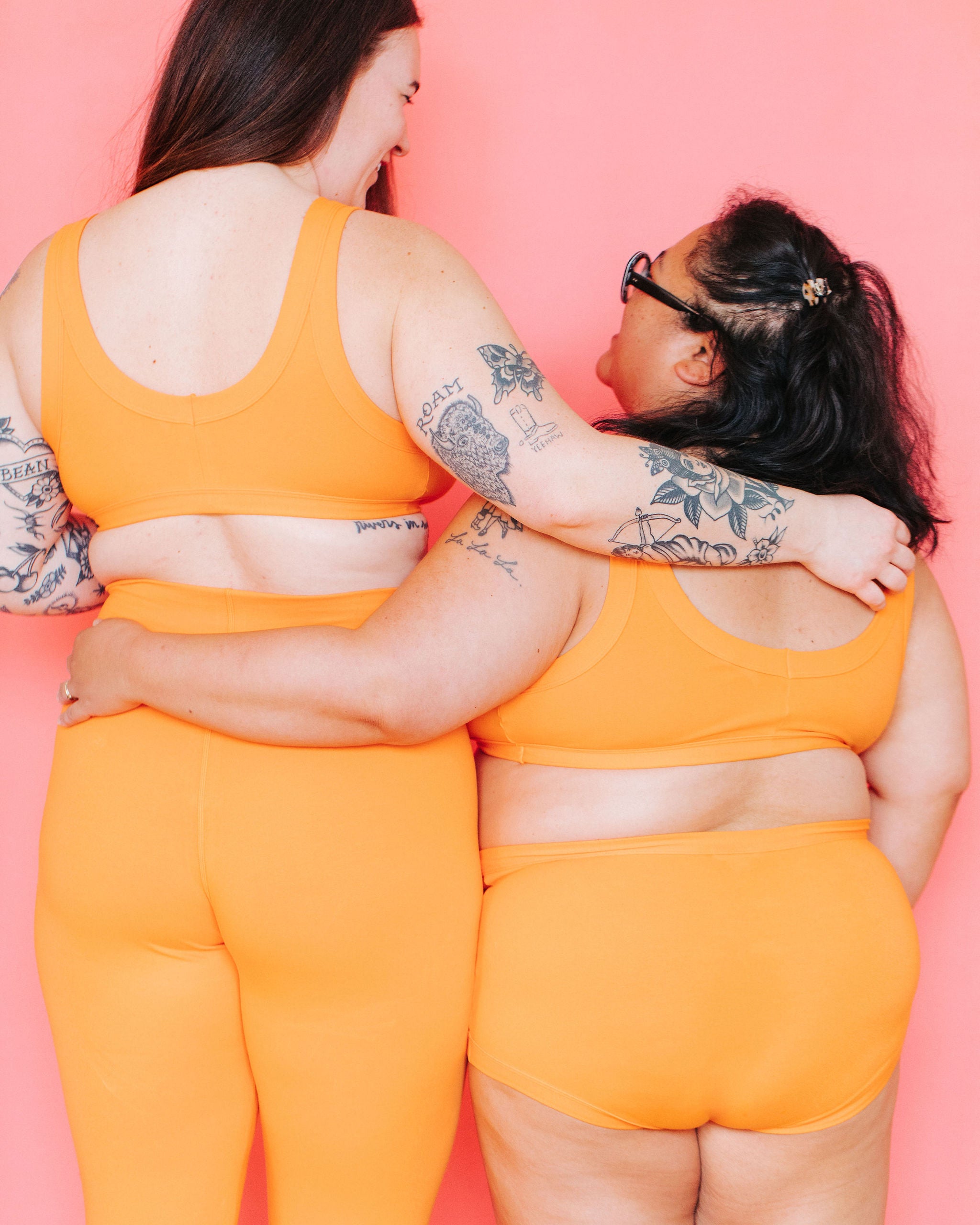 Two models with their backs to the camera in front of a pink background wearing various styles in the orange color Oregon Sunstone.