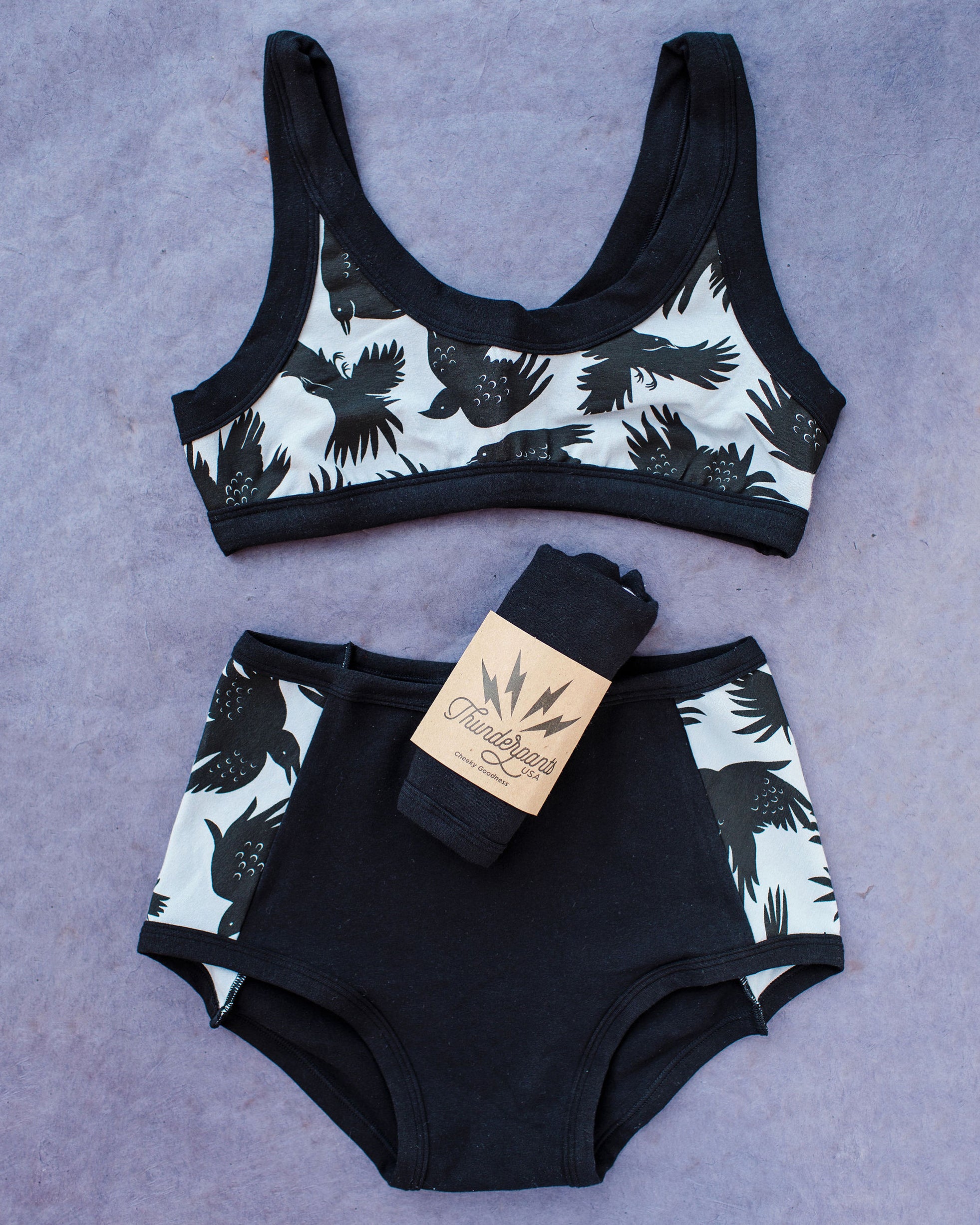 Flat lay of Thudnerpants Original Panel Pants style underwear in So it Crows - black crows with grey background one the sides and black on front and back - and Bralette in So it Crows. 