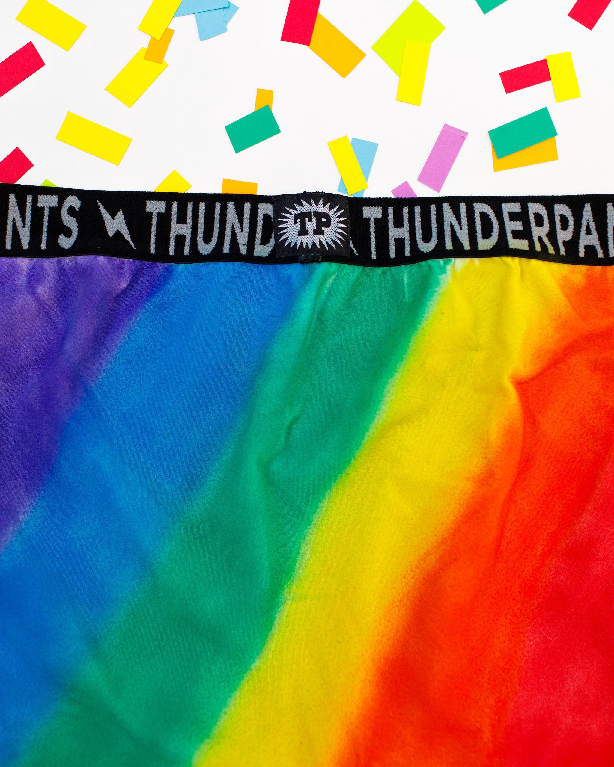 Close up of Thunderpants organic cotton Boxer Brief style underwear in hand painted rainbow stripes for Pride laying on rainbow confetti.