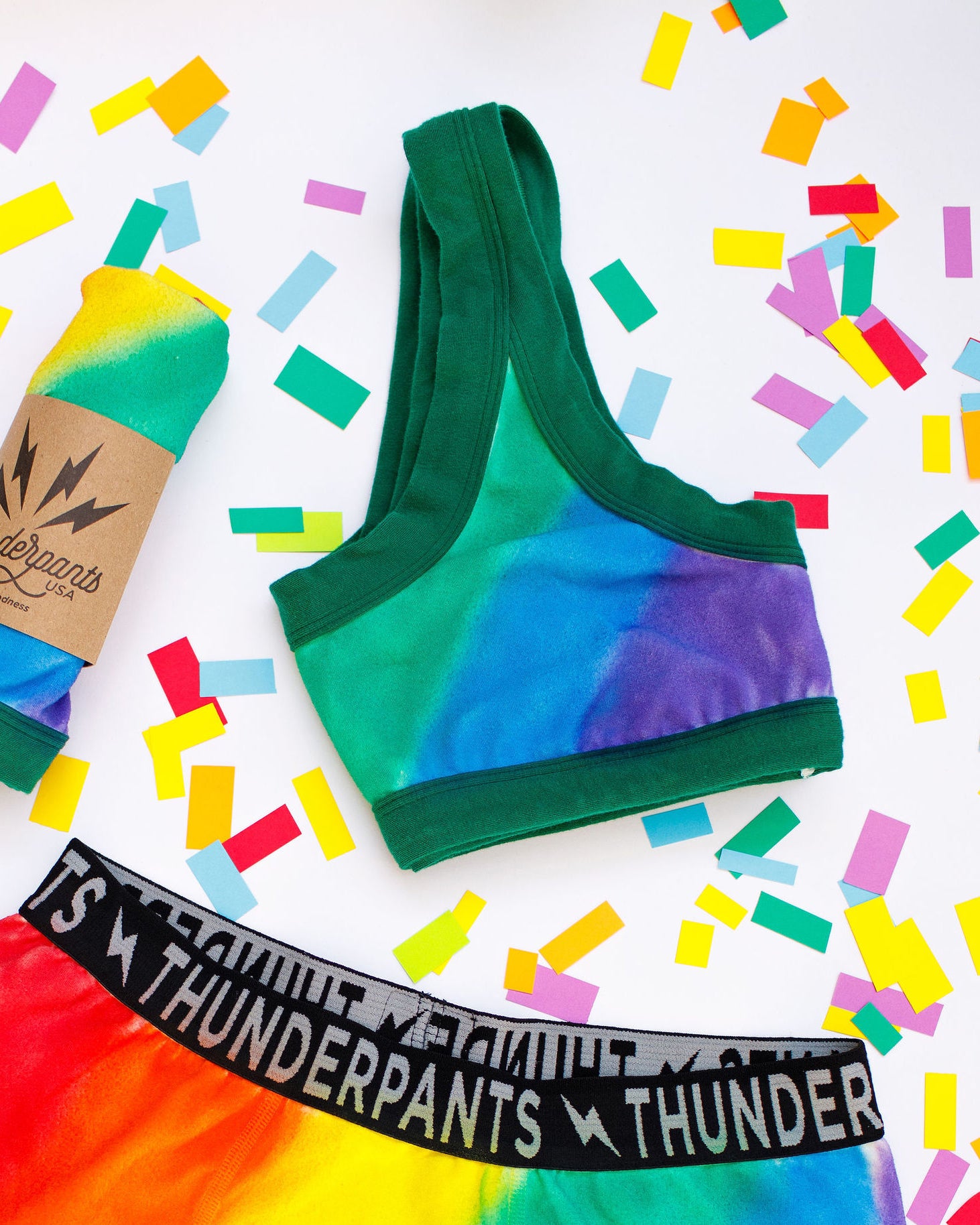Flat lay of Thunderpants organic cotton Bralette in hand painted rainbow stripes for Pride laying on confetti.