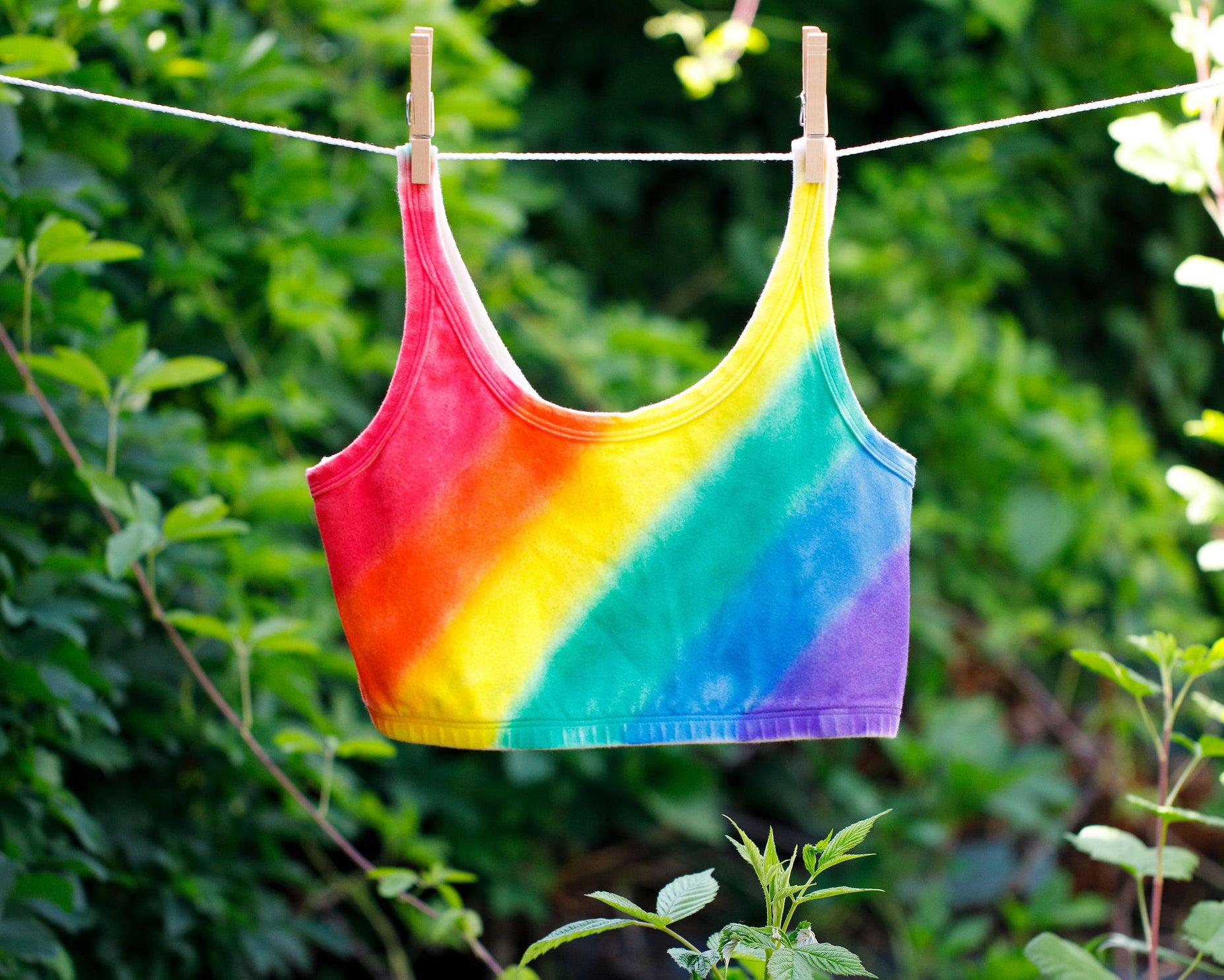 Thunderpants organic cotton Longline Bra in hand painted rainbow stripes for Pride hanging on a clothes line in greenery.