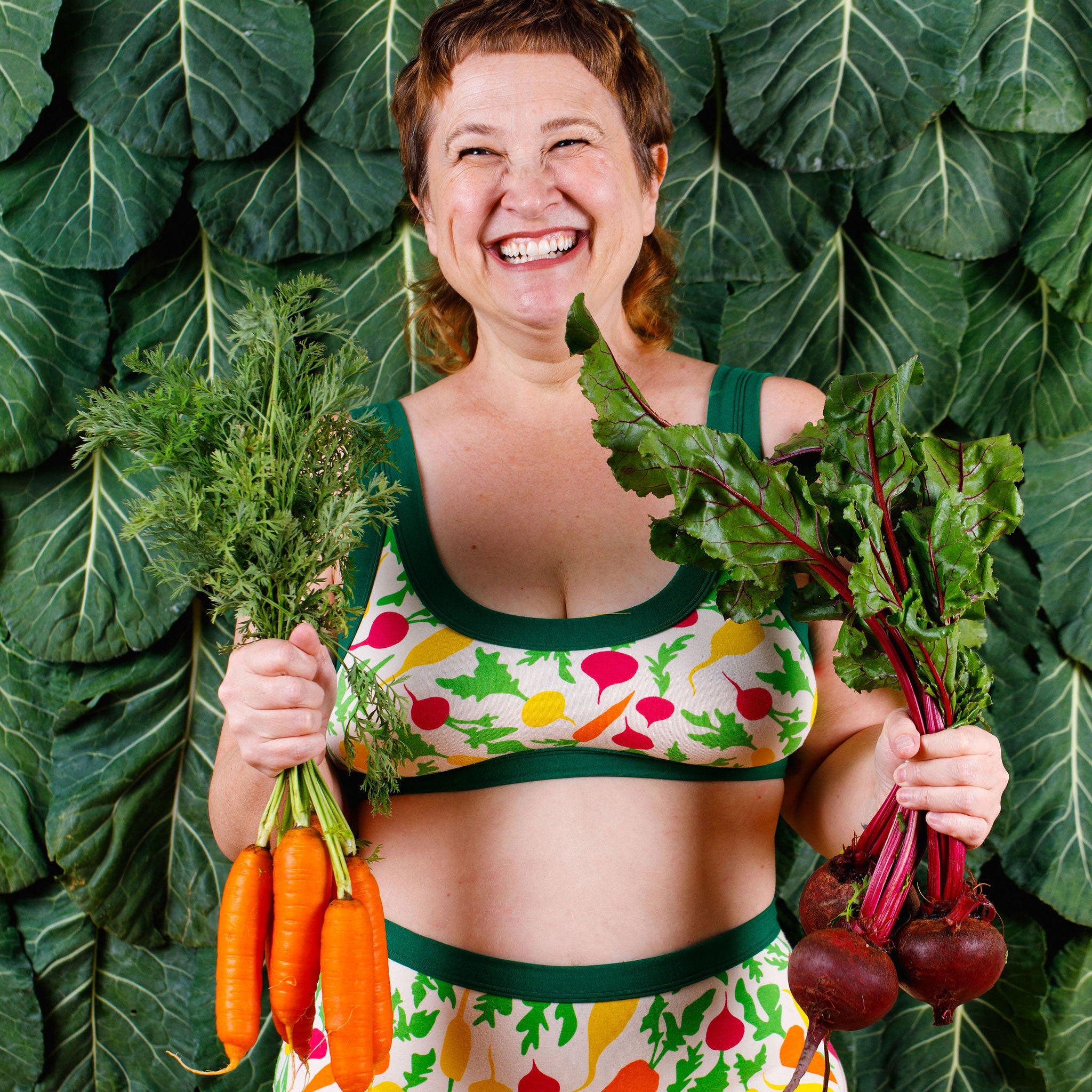 Model holding beets and carrots and smiling wearing Thunderants Sky Rise style underwear and matching Bralette in Root Veggies print - yellow, orange, pink, and green vegetables.