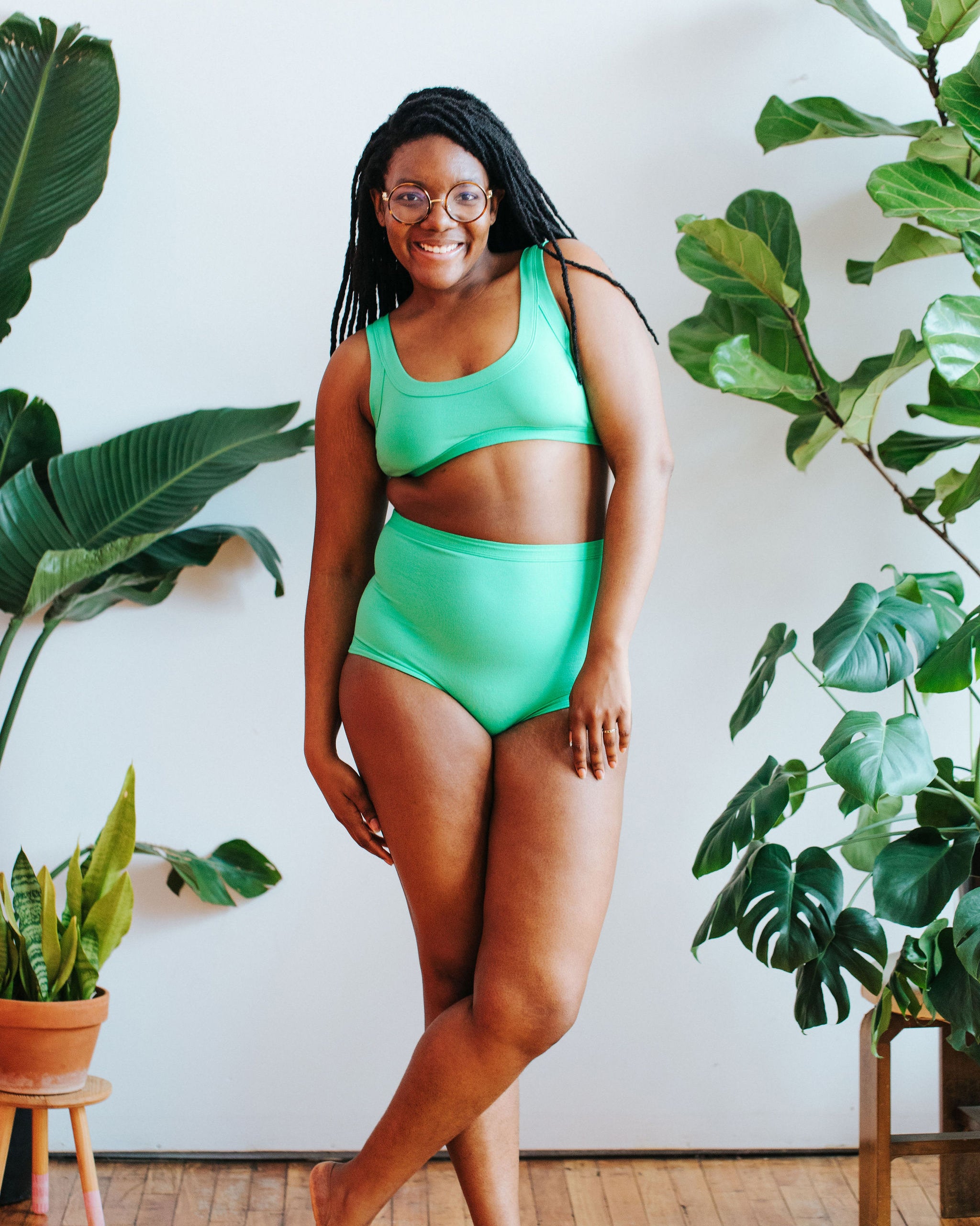 Model smiling in front of a bunch of plants wearing a Bralette and the Sky Rise style underwear in the light green color Sour Apple. 