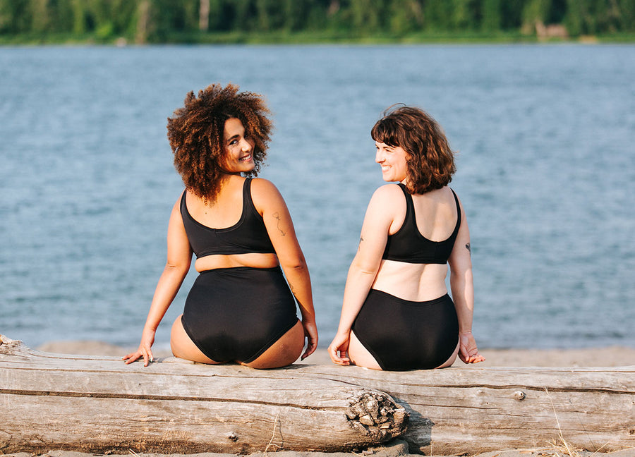 Two models sitting on a log wearing Thunderpants Swimwear sets in Black.