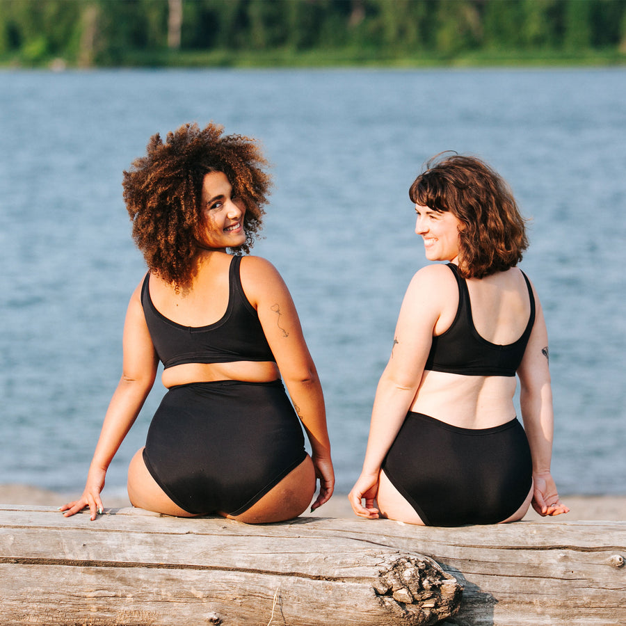 Two models sitting on a log with a river in front of them wearing Thunderpants Swimwear Sky Rise and Original bottoms and Swim Top in plain Black.